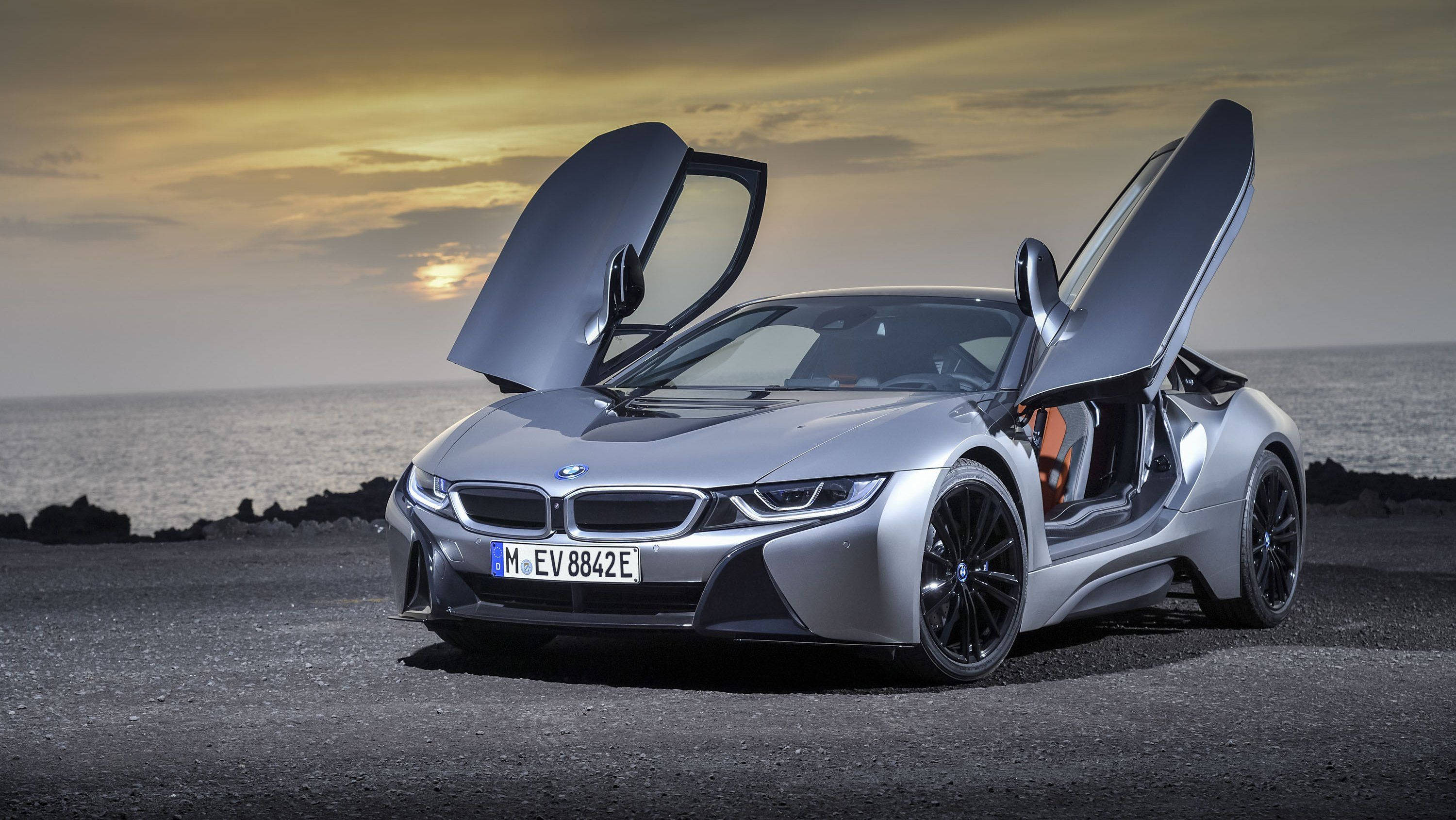BMW 動圖 桌布 壁紙 — 一架時尚的銀色BMW i8，鷗翼車門打開，停在日落時分的岩石海岸上，俯瞰海洋。