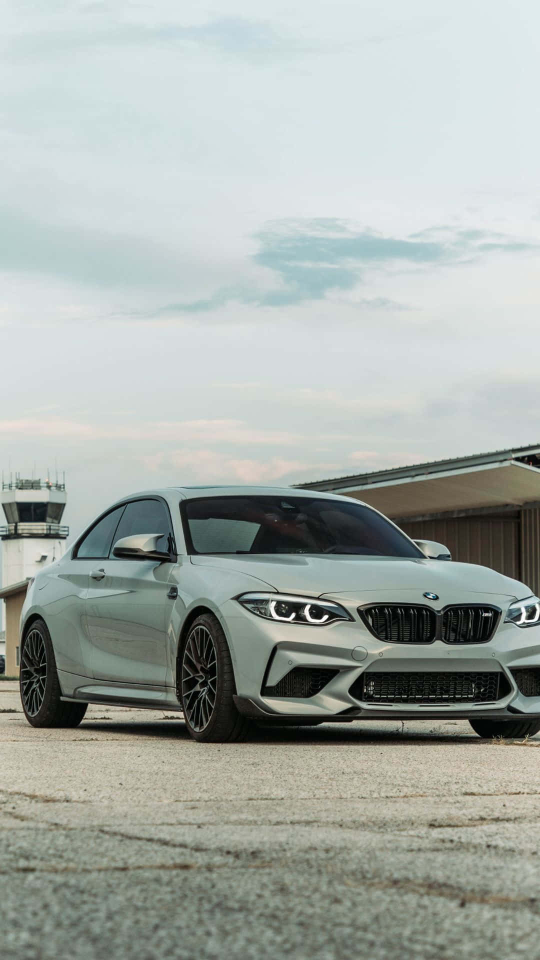 Android Bmw 桌布 壁紙 — 一架時尚的銀色BMW M2雙門跑車停放在裂開的路面上，背景是機場控制塔，天空多雲。