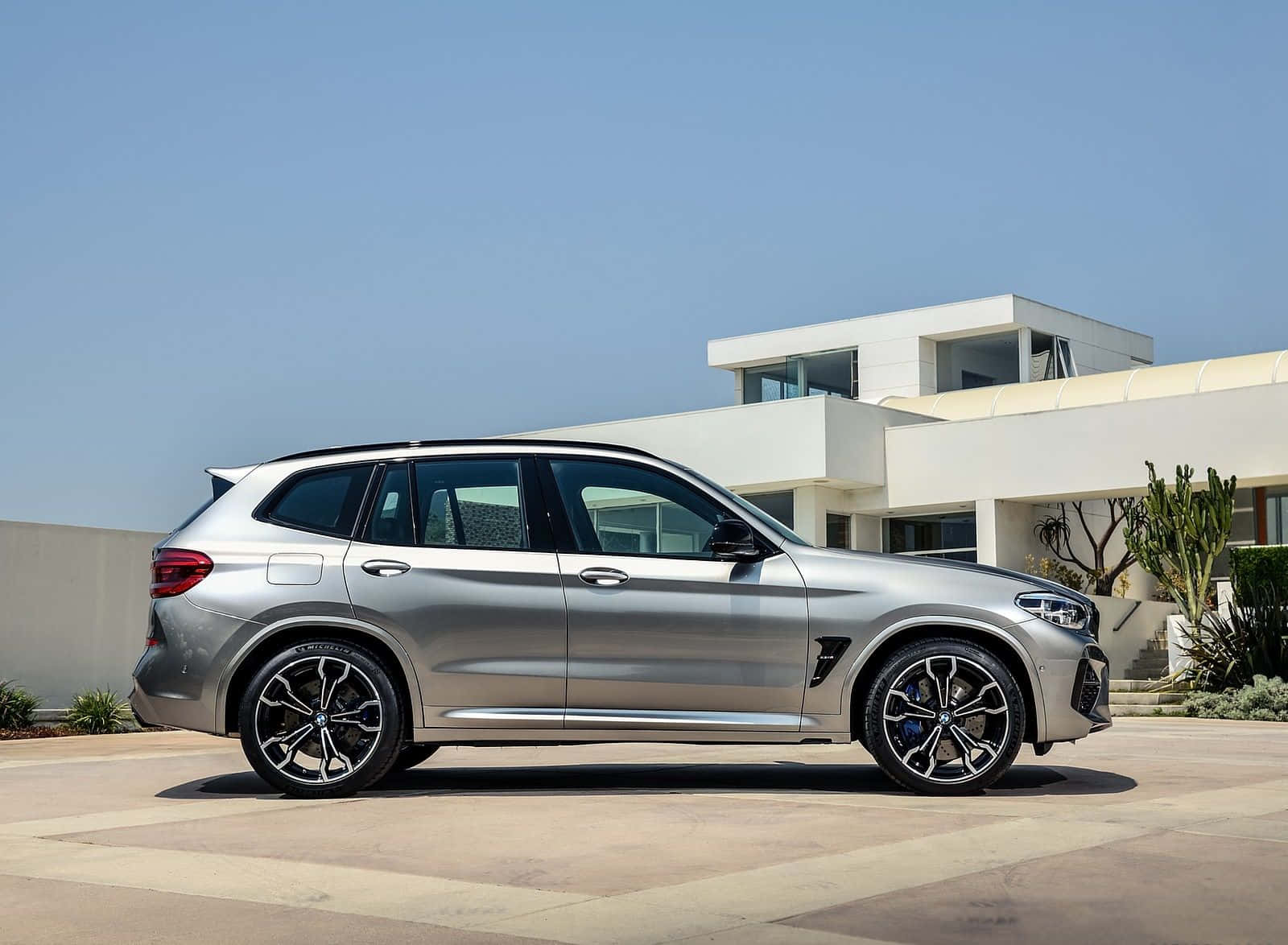 Bmw X3 桌布 壁紙 — 一架時尚的銀色BMW X5 M停放在陽光普照的混凝土車道上，前方是帶有沙漠植物的現代白色房屋。