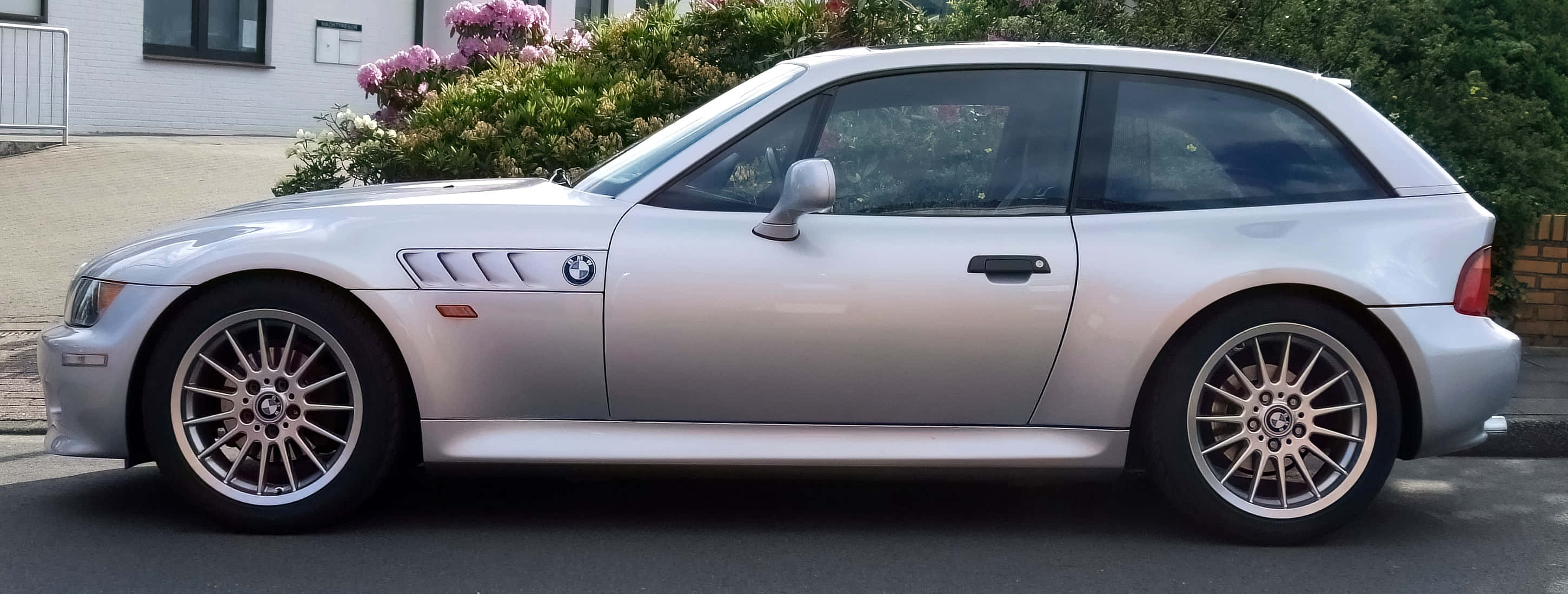 Bmw Z3 桌布 壁紙 — 一架時尚的銀色BMW Z3敞篷跑車停放在住宅街道上，背景有盛開的粉紅色花朵和綠色灌木。