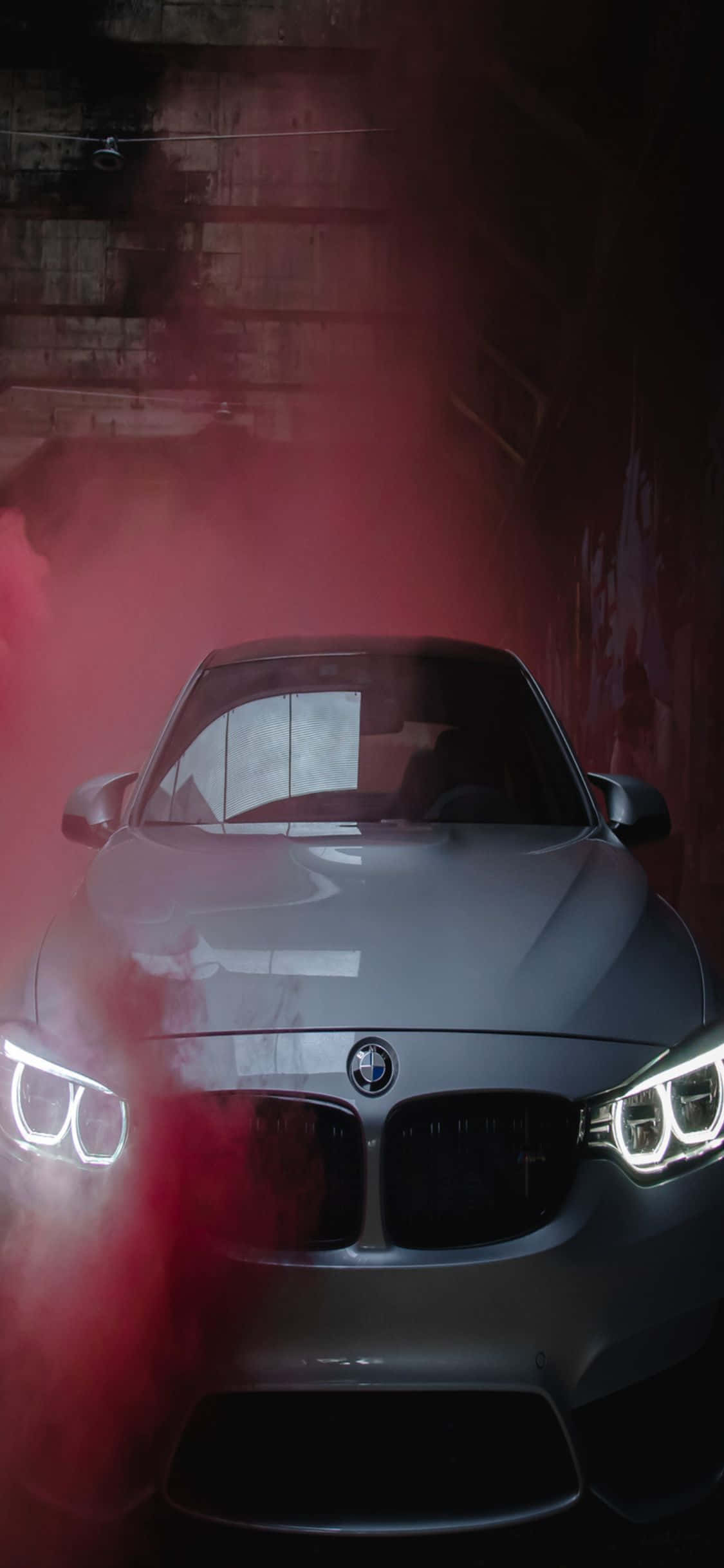 iPhone X Bmw 桌布 壁紙 — 一架時尚的銀色BMW跑車在昏暗的混凝土隧道中，透過旋轉的紅色煙霧發出明亮的車頭燈光芒。