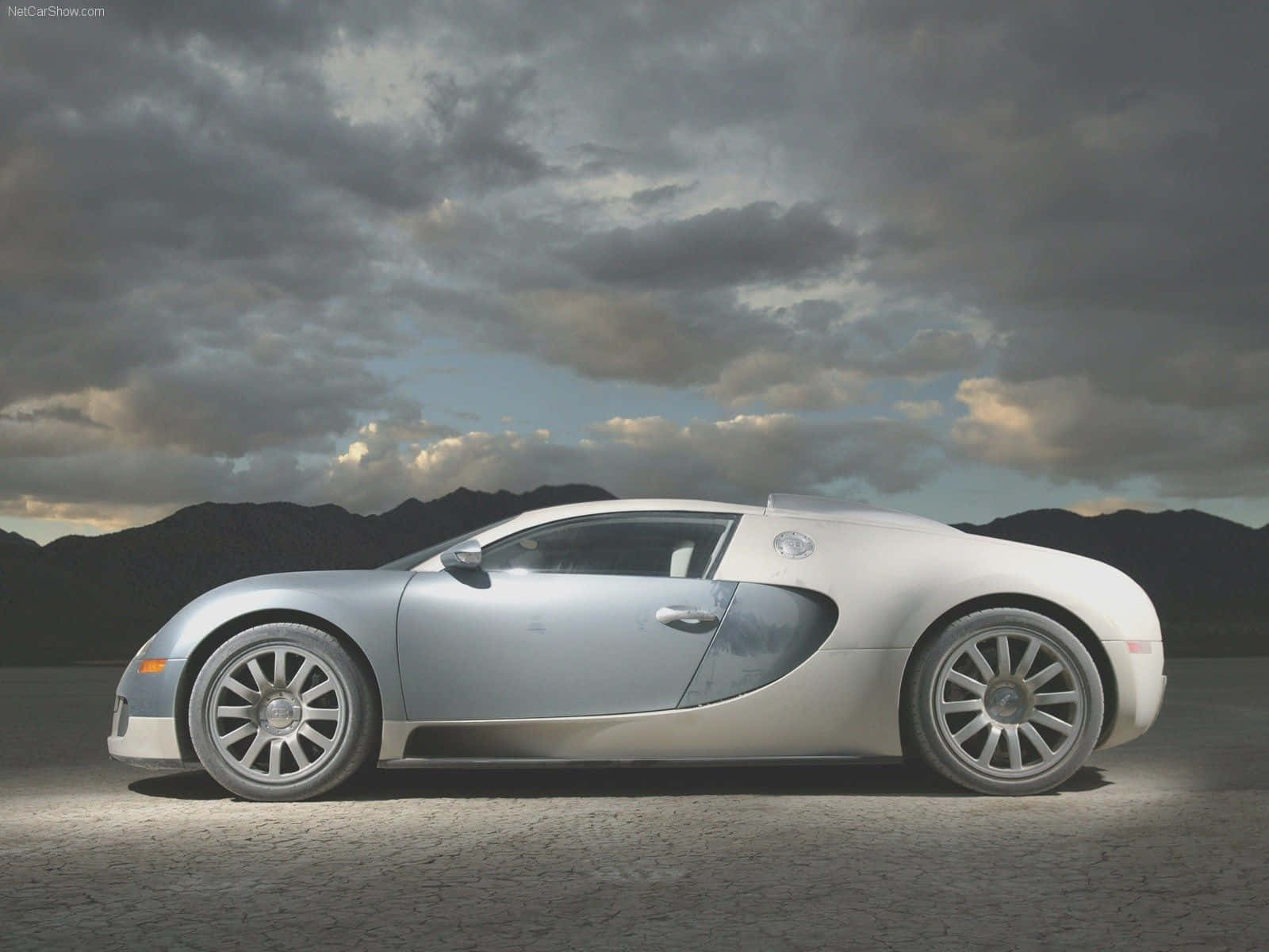 Best Bugatti 桌布 壁紙 — 一架時尚的銀色Bugatti跑車停泊在龜裂的土地上，背景是戲劇性的多雲天空和遠山。