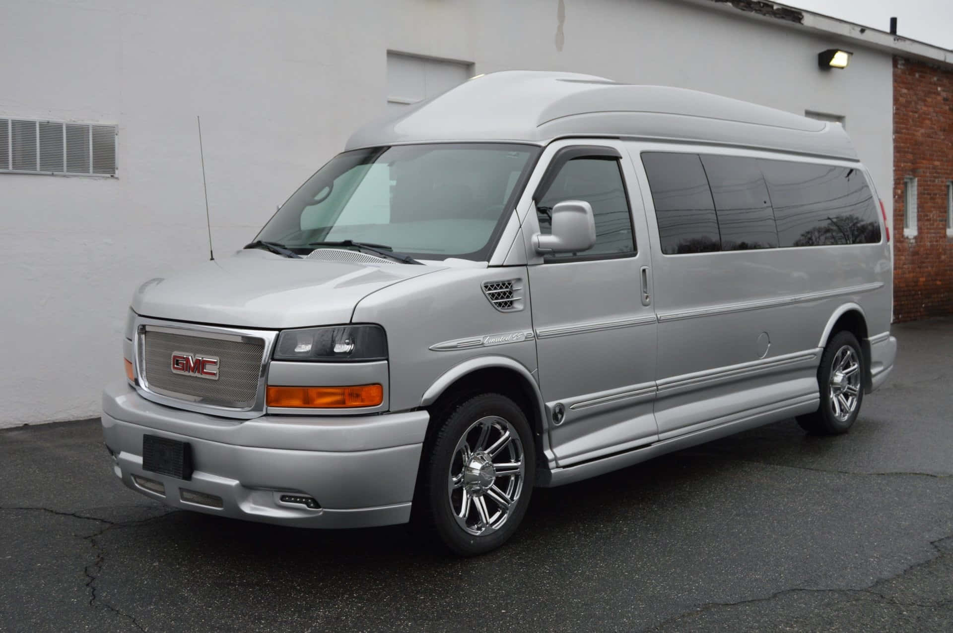 Gmc Savana 桌布 壁紙 — 一架時尚的銀色GMC露營車停泊在瀝青路上，旁邊是白色磚砌建築，天空多雲。