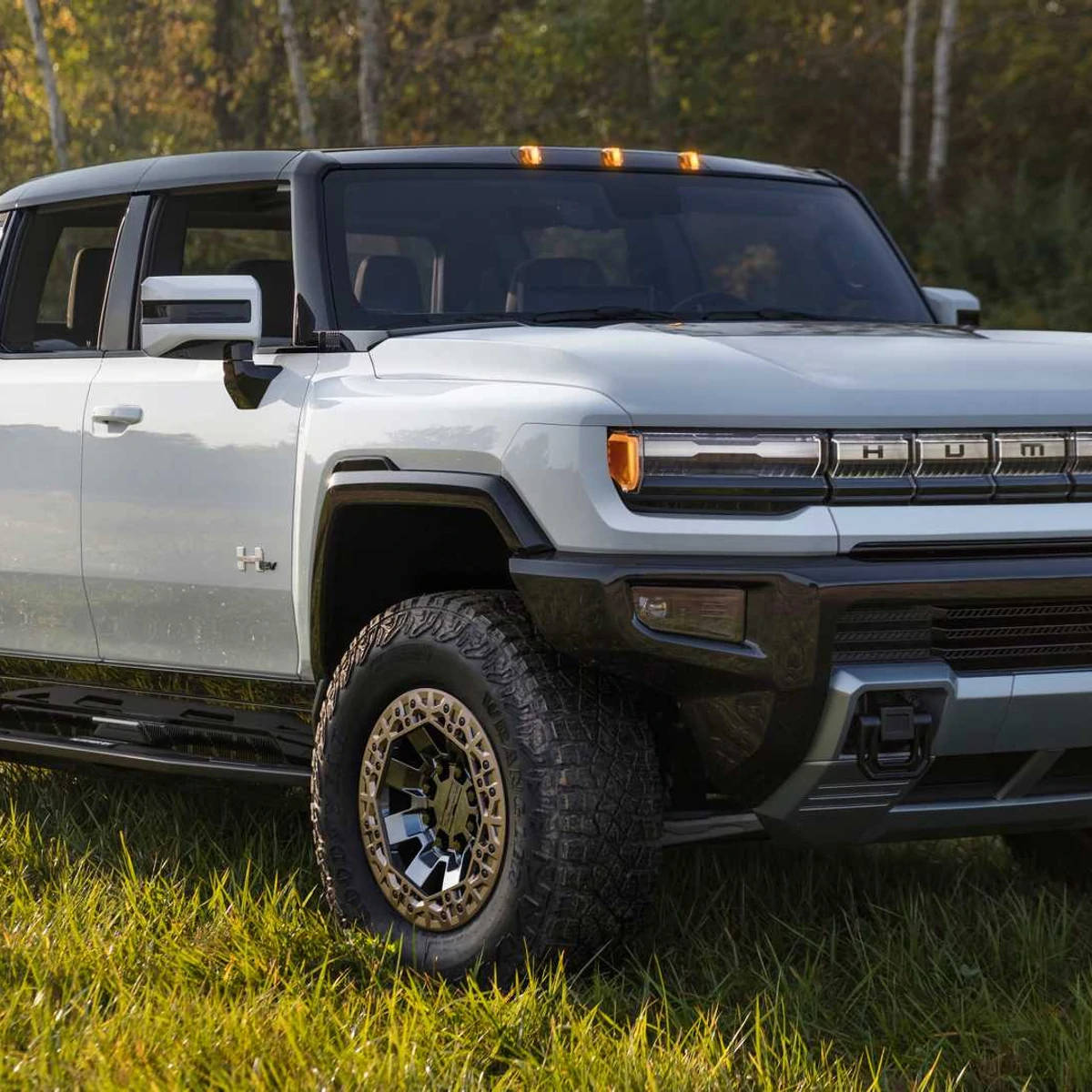 Truck 桌布 壁紙 — 一架時尚的銀色Hummer EV在金色陽光下，背後有森林樹木，高高矗立在草地上。
