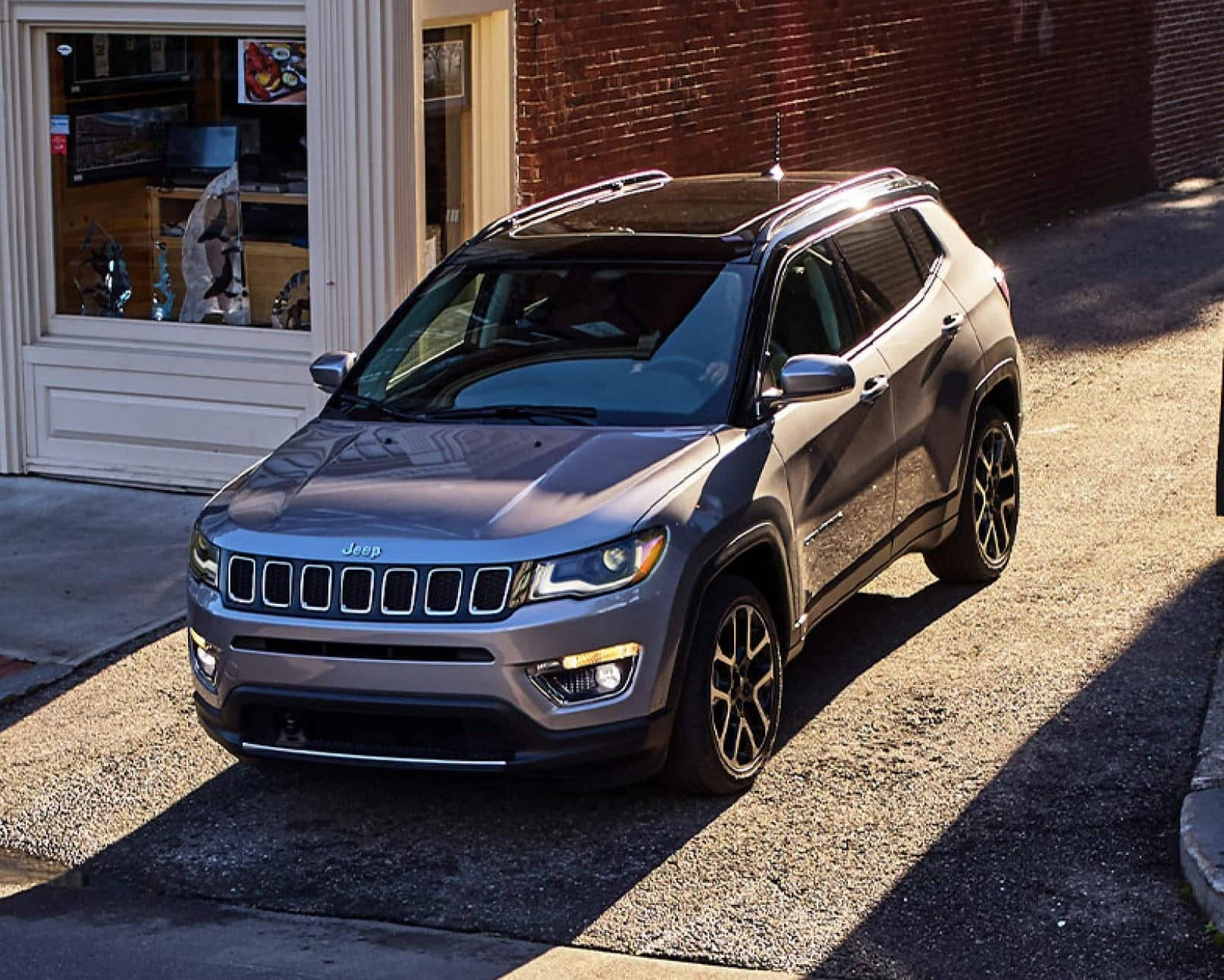 汽車 桌布 壁紙 — 一架時尚的銀色Jeep Compass停泊在陽光普照的城市街道上，旁邊是磚砌建築和商店門面。