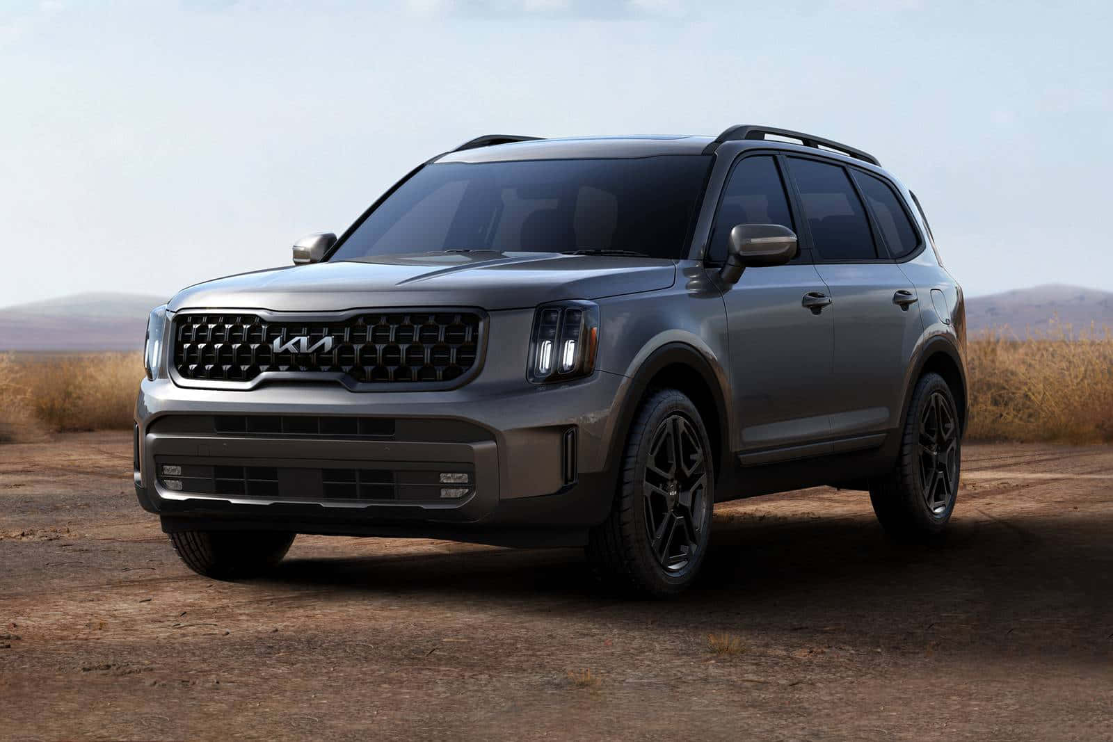 Kia Telluride 桌布 壁紙 — 一架時尚的銀色Kia SUV停泊在塵土飛揚的沙漠道路上，乾草和遠山在朦朧的天空下。