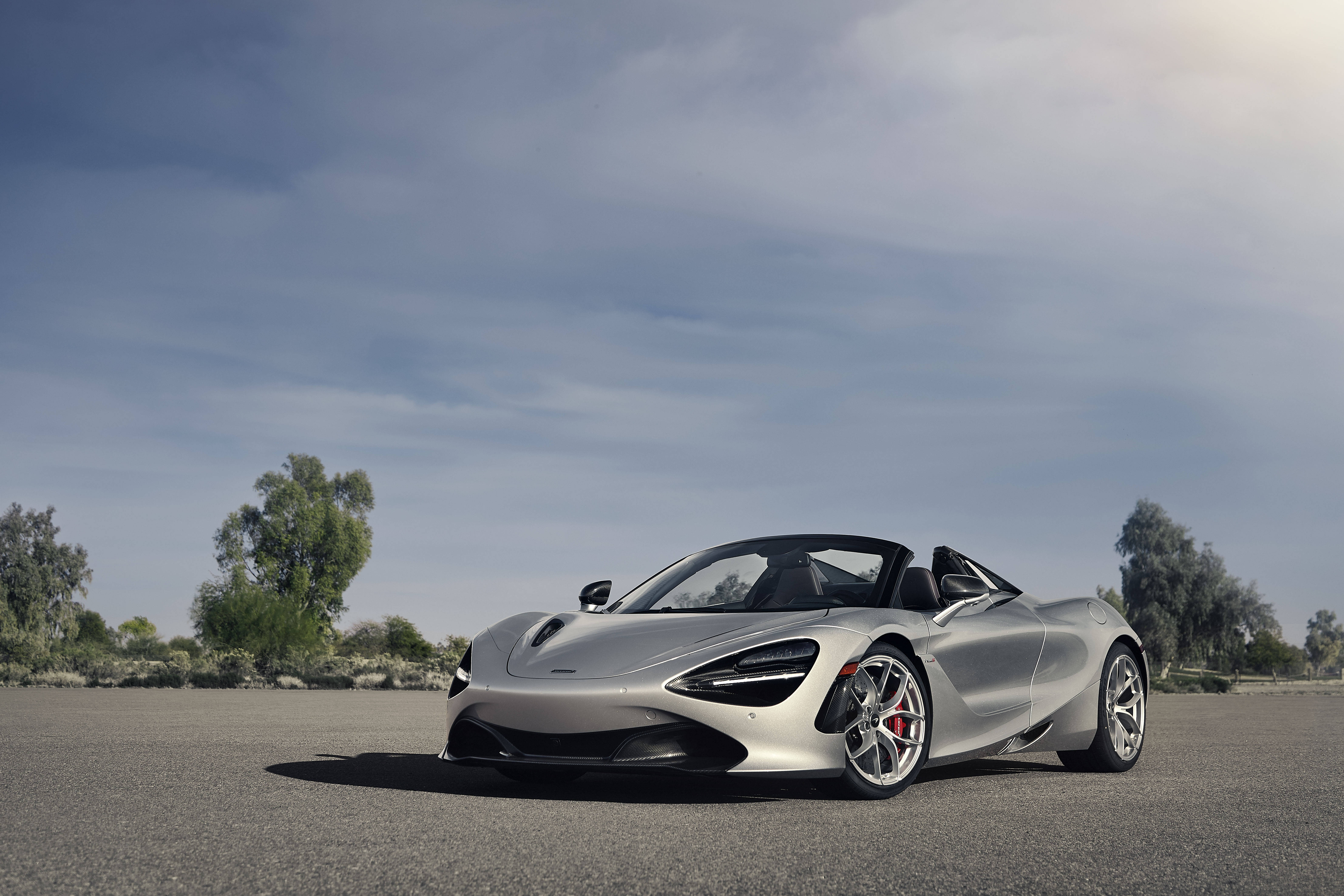 麥拉倫 720S 桌布 壁紙 — 一架時尚的銀色McLaren 720S Spider敞篷跑車停放在開闊的柏油路上，天空部分多雲，背景有稀疏的樹木。