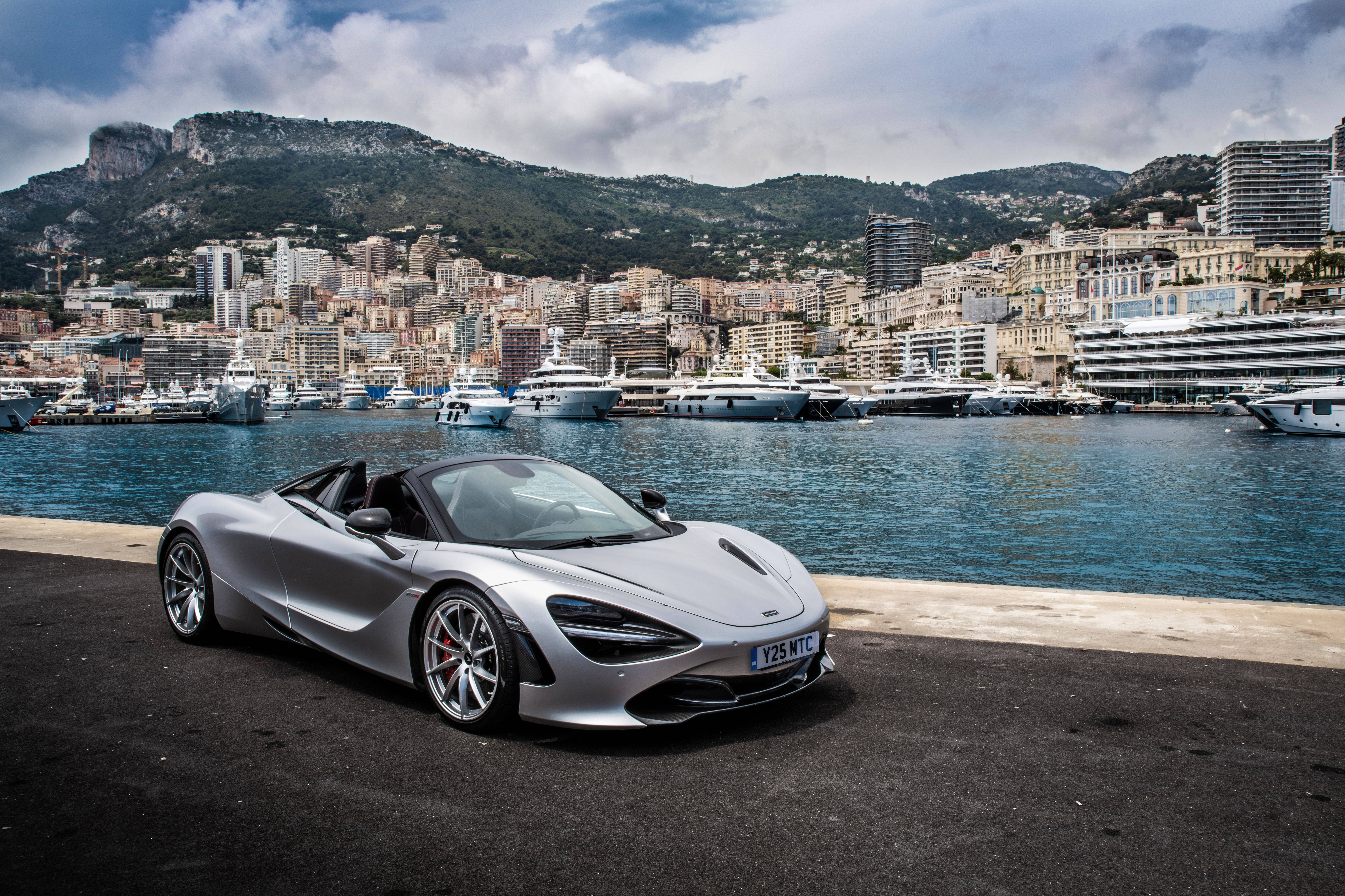 Mclaren 720s 桌布 壁紙 — 一架時尚的銀色McLaren 720S Spider停在海濱長廊上，背景是豪華遊艇和密集的沿海城市景觀。