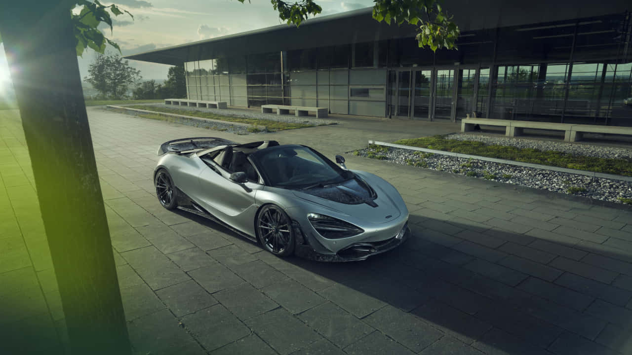 汽車 桌布 壁紙 — 一架時尚的銀色McLaren Speedtail敞篷跑車停放在現代玻璃建築旁的石頭廣場上，陽光穿過樹葉灑落。
