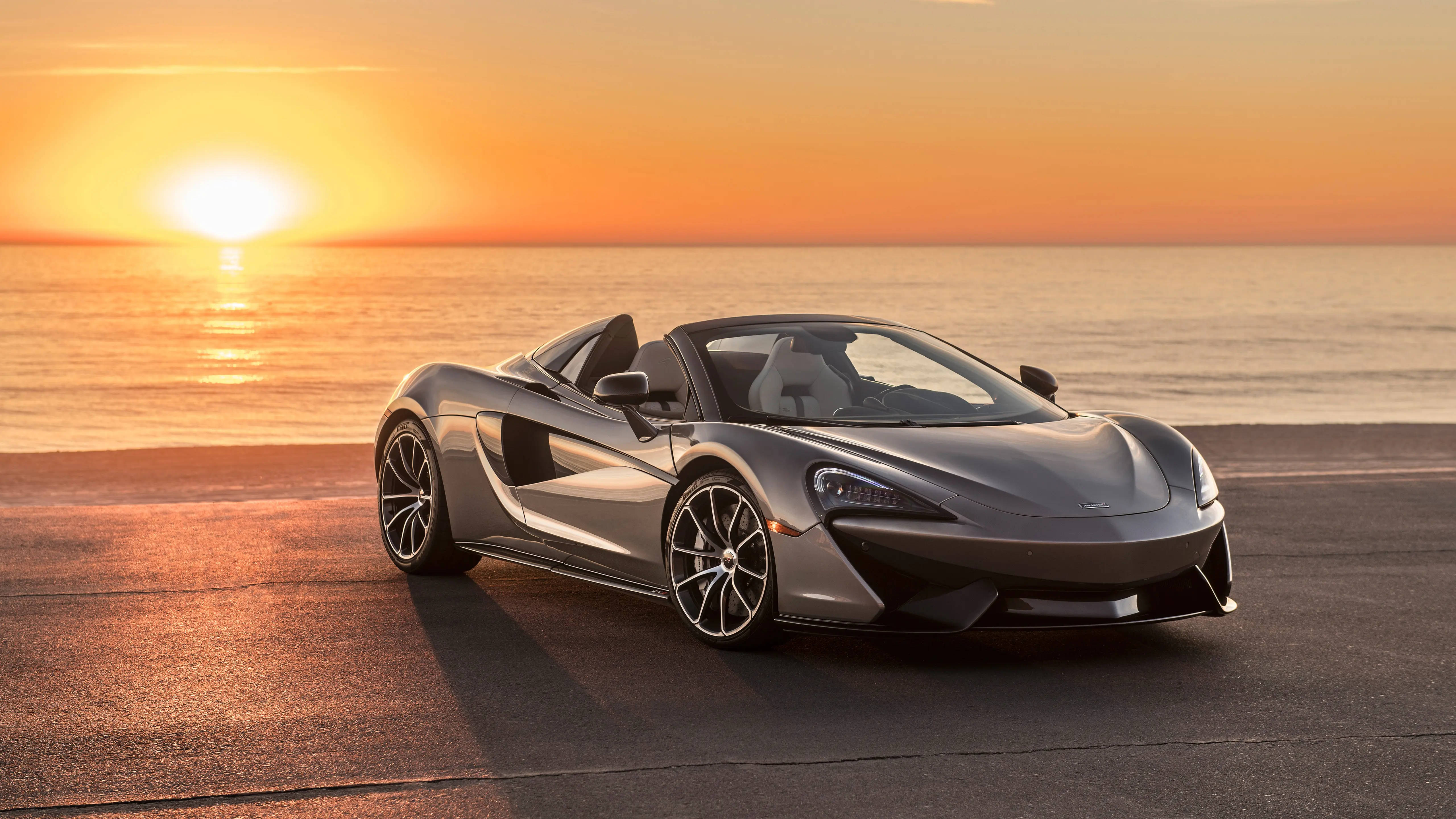 跑車 桌布 壁紙 — 一架時尚的銀色McLaren跑車停泊在海邊道路上，夕陽在海面上落下，投射出溫暖的橙色光芒。