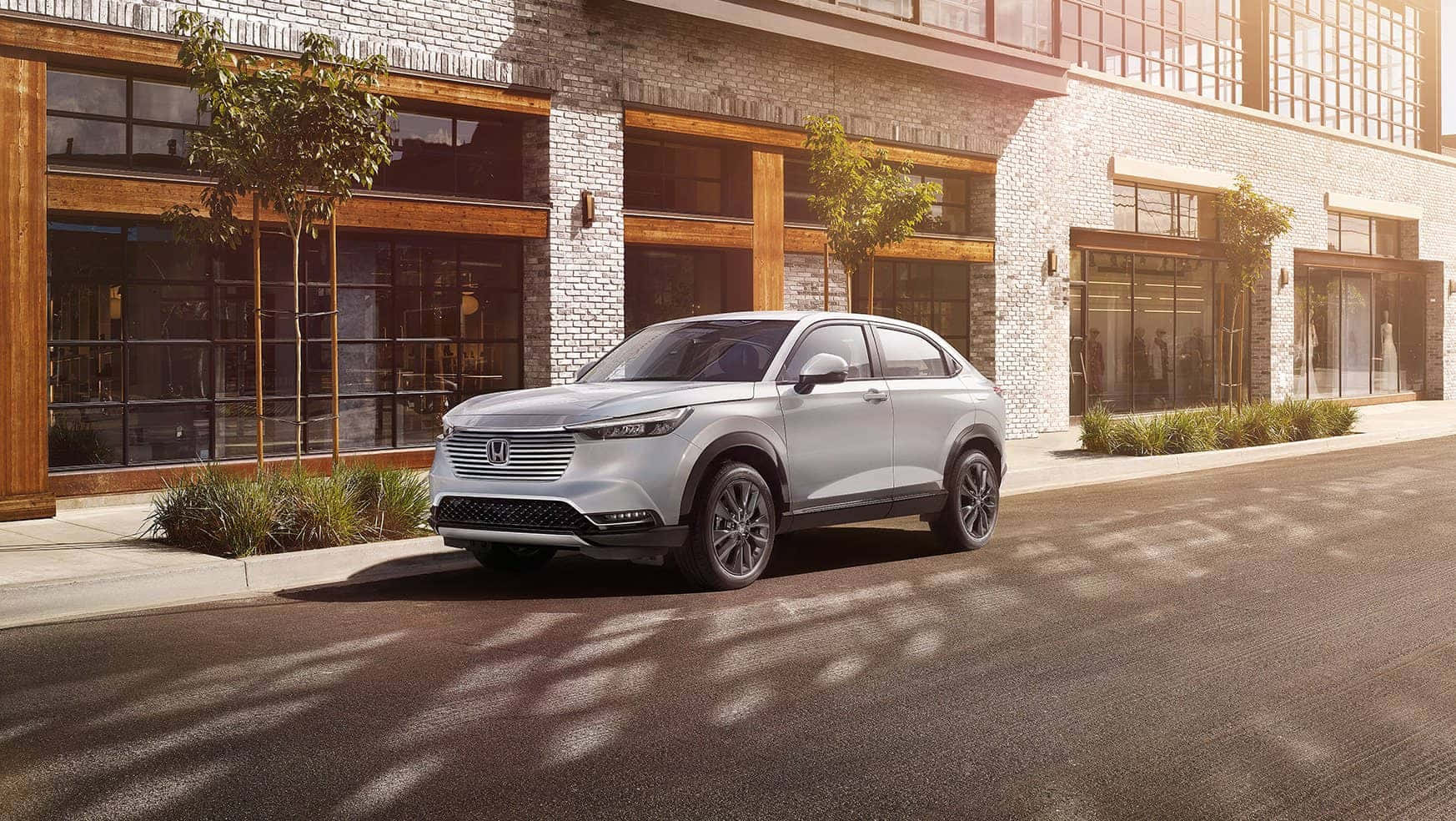 Honda Hr-v 桌布 壁紙 — 一架時尚的銀色SUV停放在陽光普照的城市街道上，旁邊是帶有大型玻璃窗的現代磚砌建築。