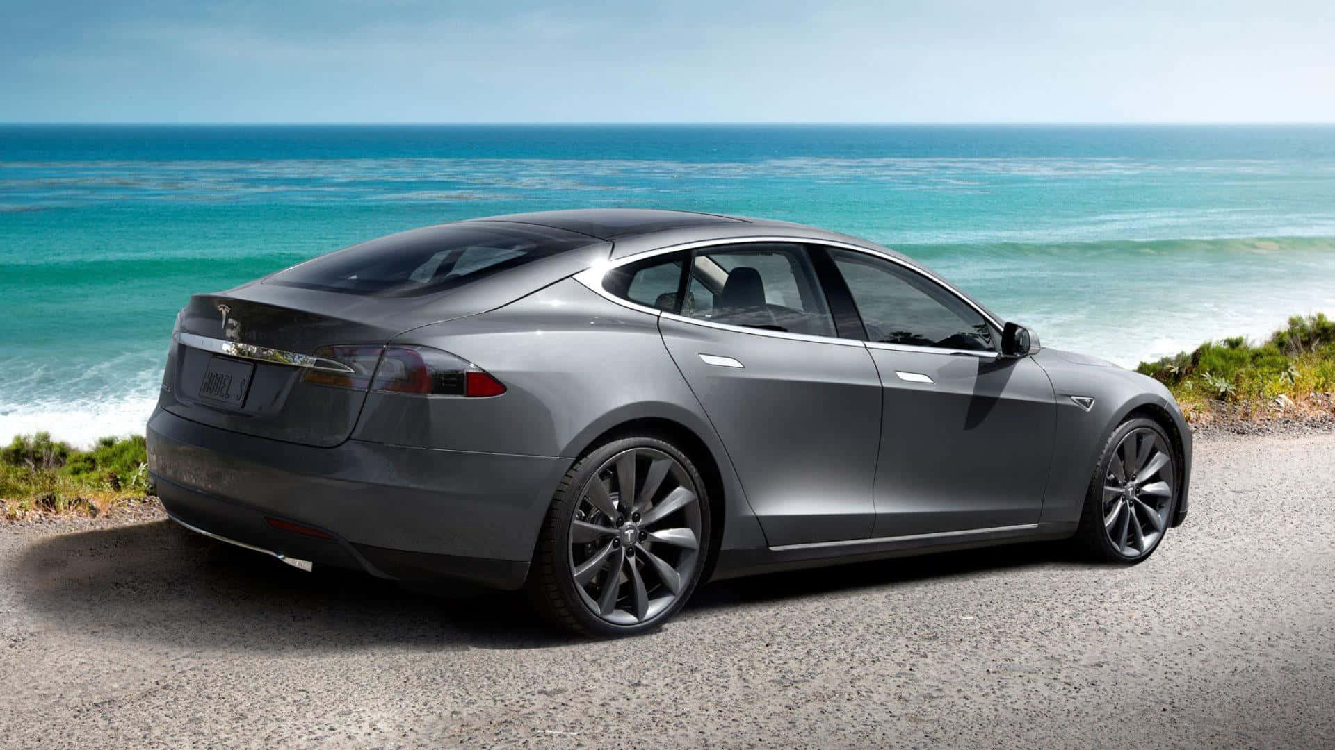 Tesla Model 3 桌布 壁紙 — 一架時尚的銀色Tesla Model S停泊在碎石路上，俯瞰著清澈藍天下碧綠色的海浪。