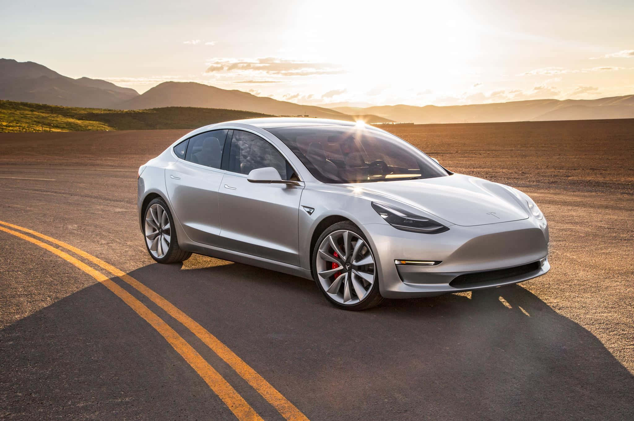 Tesla Model 3 桌布 壁紙 — 一架時尚的銀色Tesla Model 3停在沙漠公路上，背景是金色的日落和山脈。
