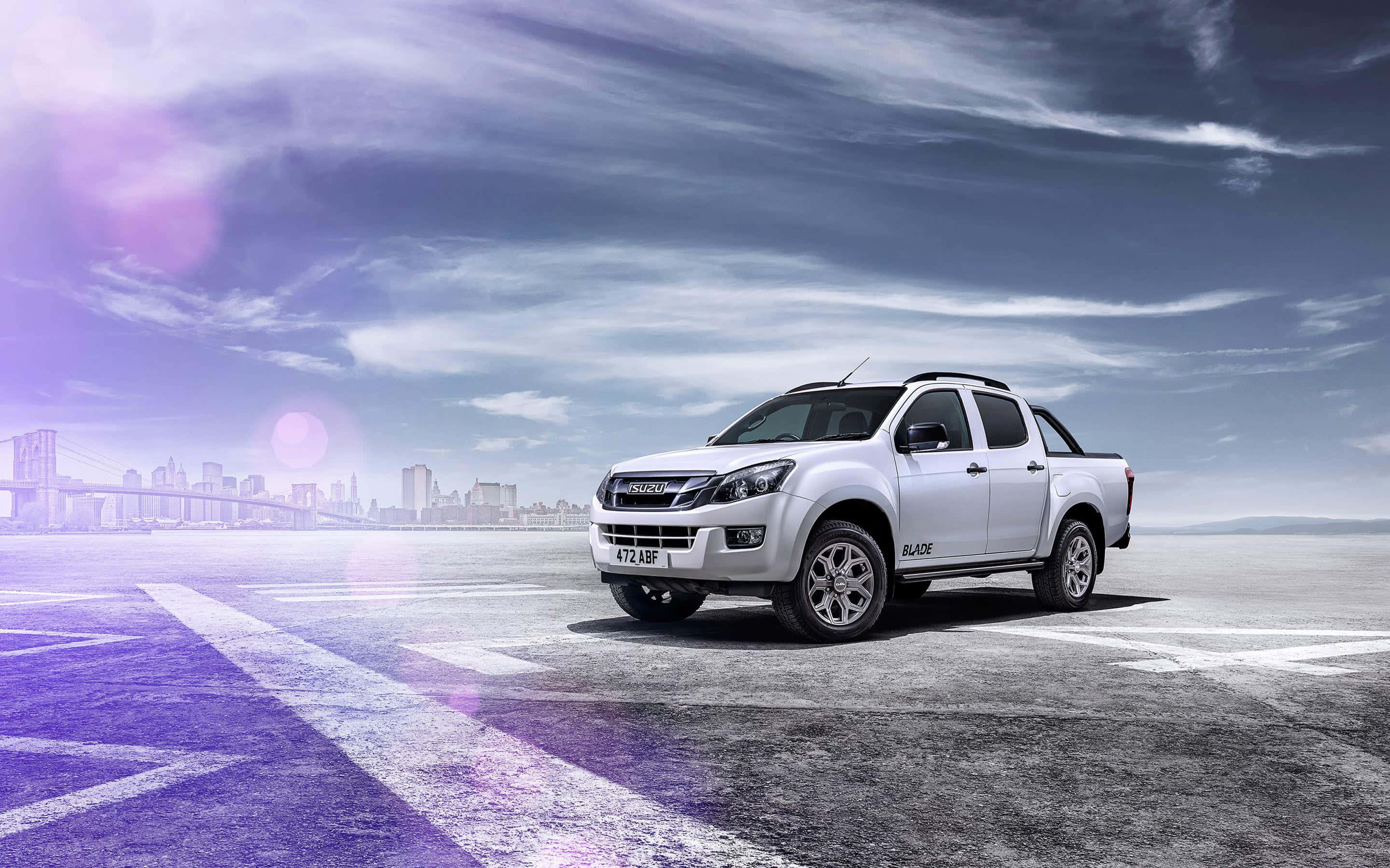 Isuzu 桌布 壁紙 — 一架時尚的銀色五十鈴皮卡停放在廣闊的鋪砌區域，背景是城市天際線和橋樑，天空呈現戲劇性的紫色和藍色。