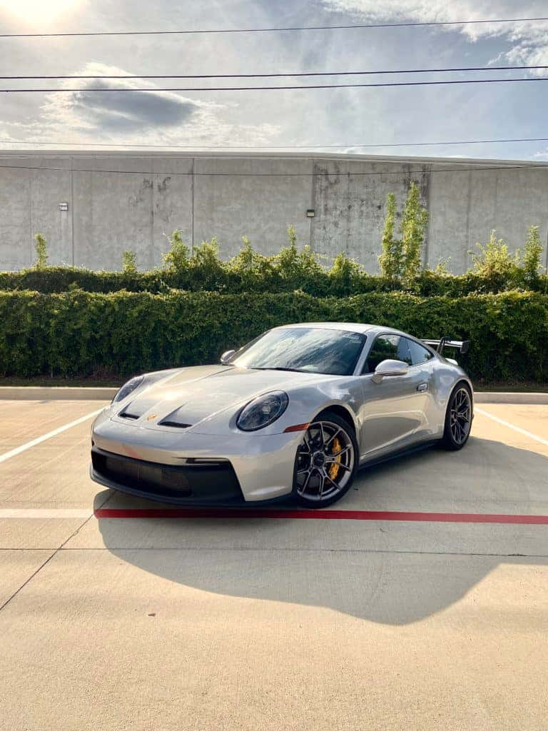 768x1024 桌布 壁紙 — 一架時尚的銀色保時捷911 GT3停放在陽光照射的混凝土停車場，後面有綠色樹籬和灰色牆壁。