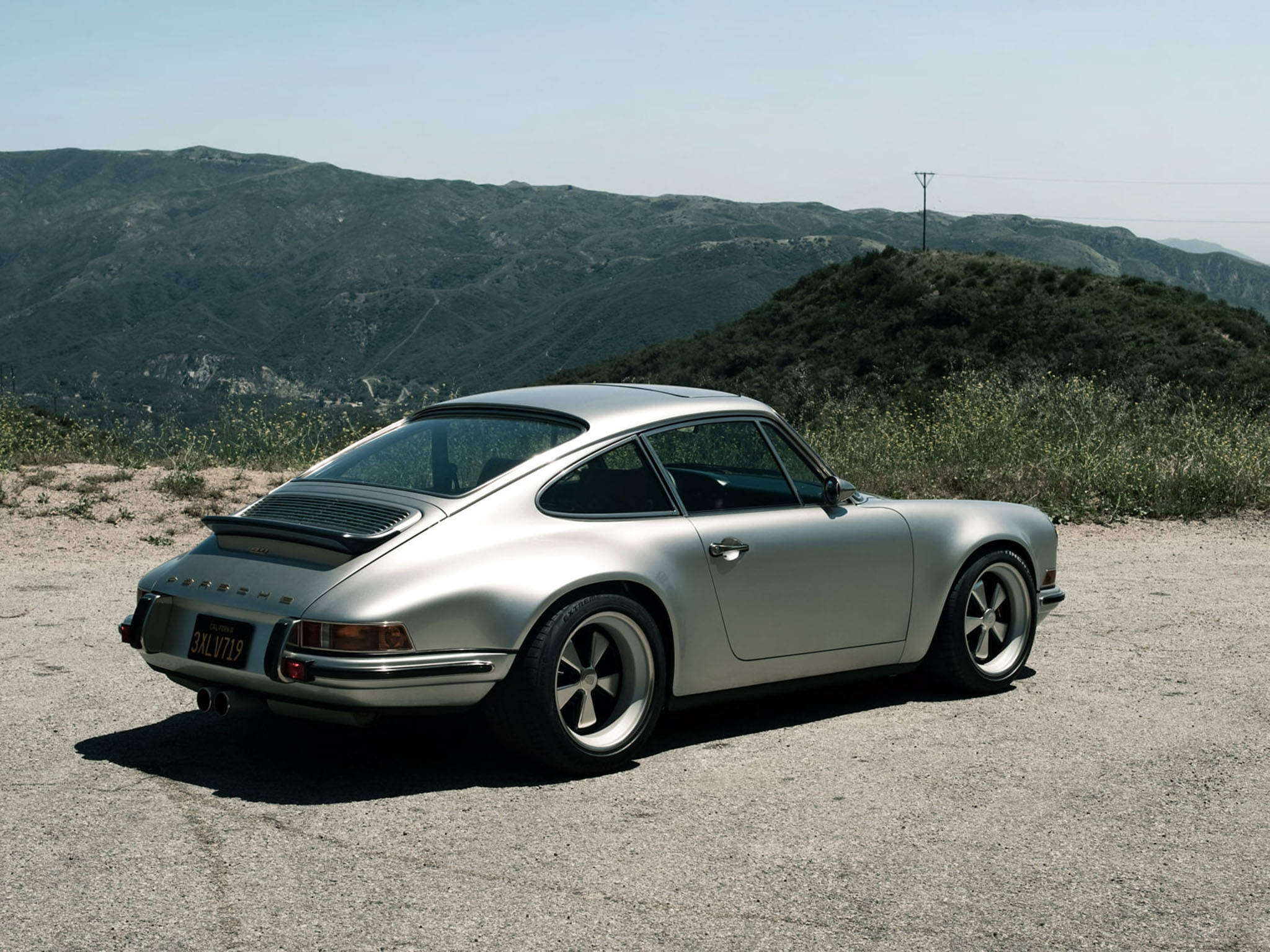 Singer 桌布 壁紙 — 一架時尚的銀色保時捷911經典跑車停泊在塵土飛揚的路邊，背景是連綿起伏的綠色山丘，頭頂是清澈的藍天。
