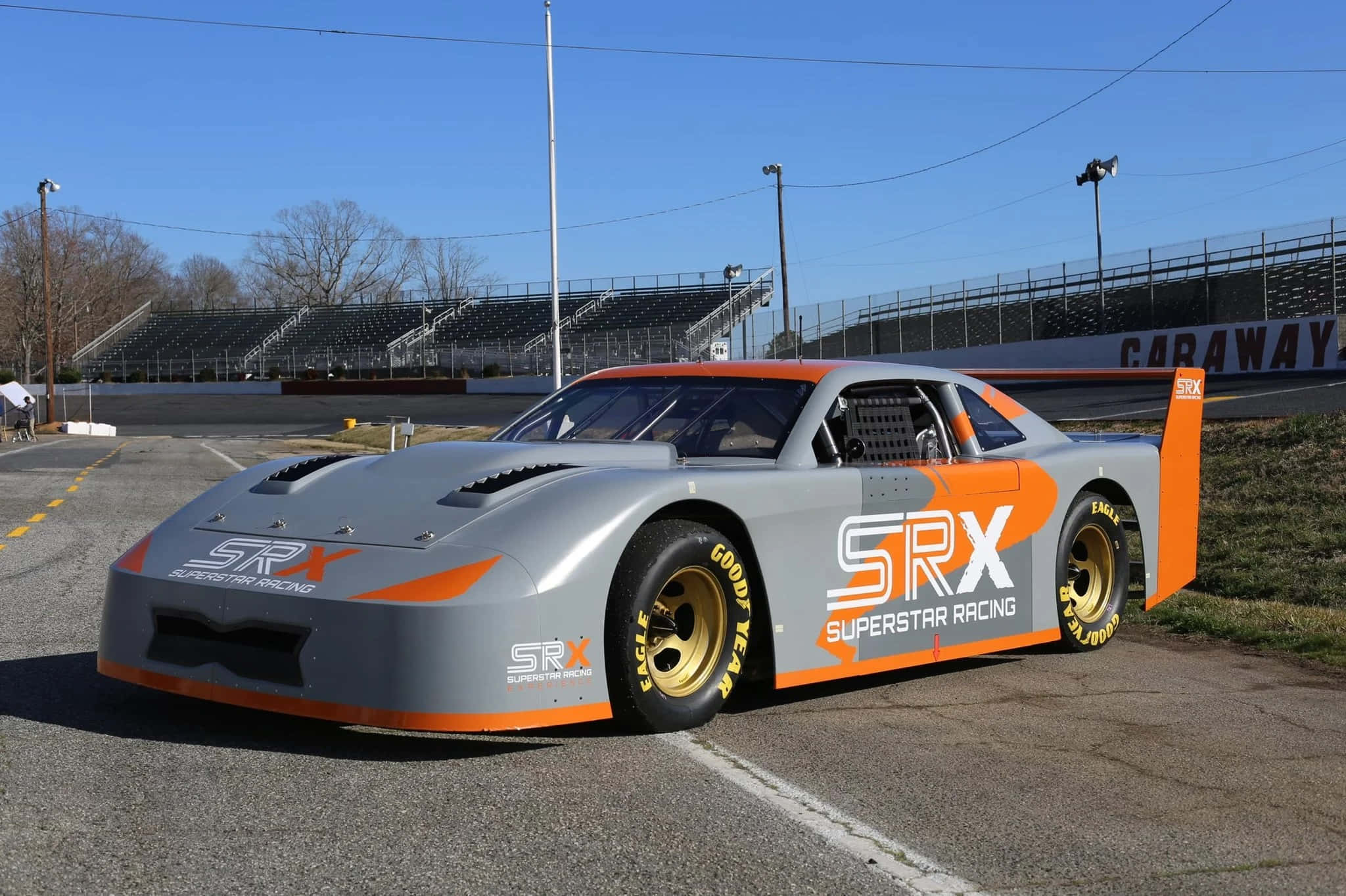 Car Racing 桌布 壁紙 — 一架時尚的銀色和橙色 SRX Superstar 賽車停泊在賽道上，背景是晴朗的藍天和空無一人的看台。