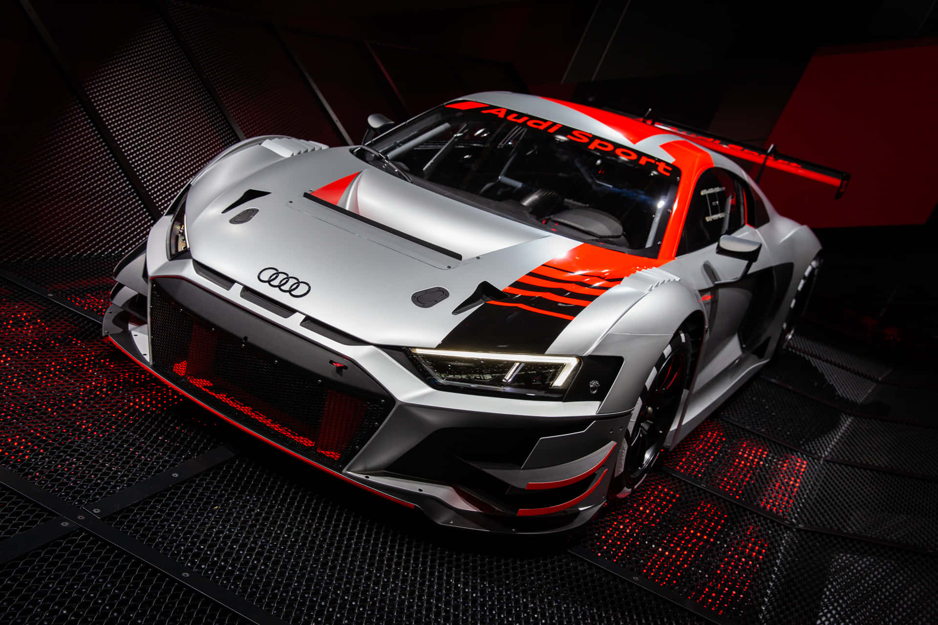奧迪 桌布 壁紙 — 一架時尚的銀色奧迪 R8 LMS GT3 賽車，帶有鮮明的紅色點綴和發光的底盤燈，停在光線昏暗的車庫中的黑色穿孔地板上。