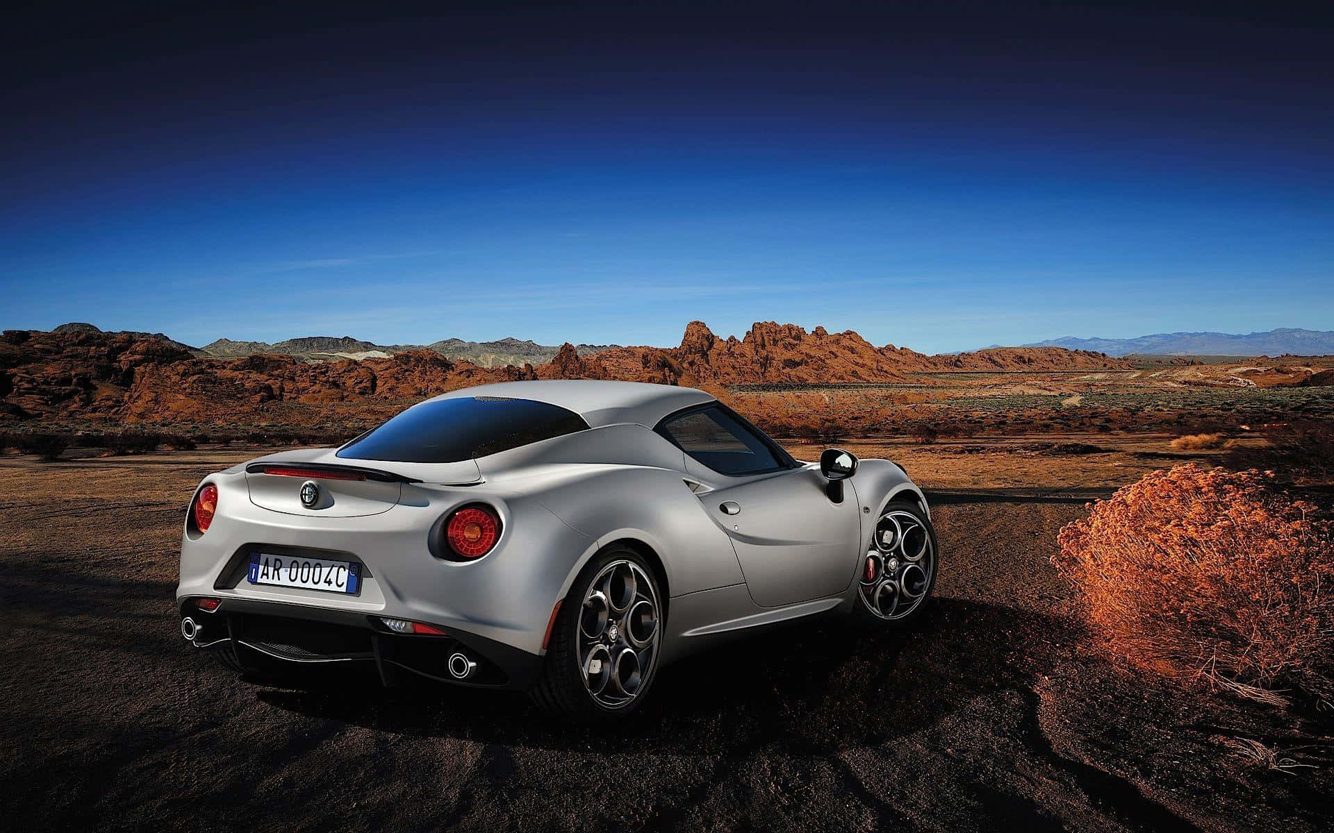 Alfa Romeo 4c 桌布 壁紙 — 一架時尚的銀色愛快羅密歐跑車停泊在崎嶇的沙漠地形上，天空清澈湛藍。