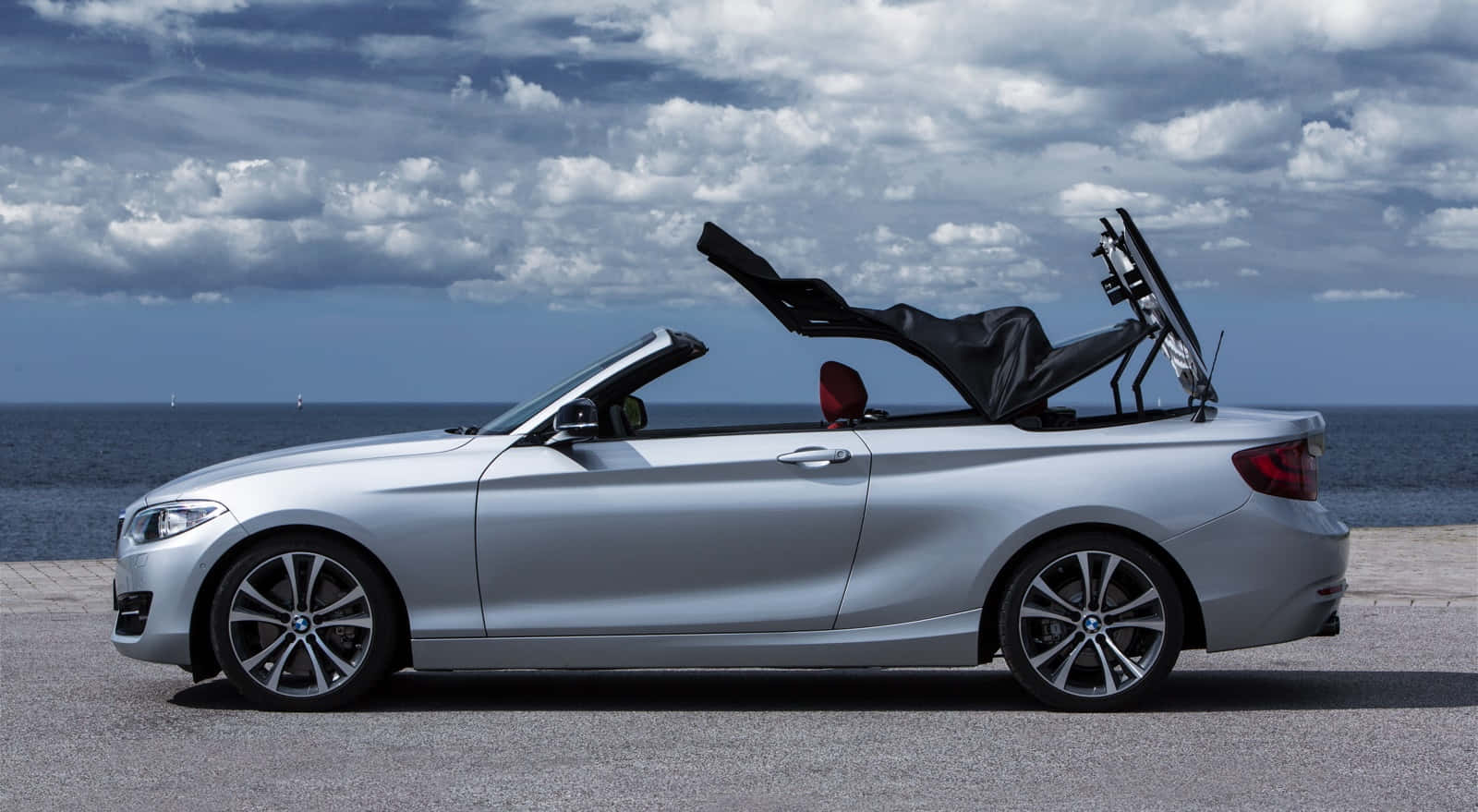 Bmw 2 Series 桌布 壁紙 — 一架時尚的銀色敞篷跑車，車頂敞開，停泊在海洋旁邊，天空佈滿戲劇性的雲層。