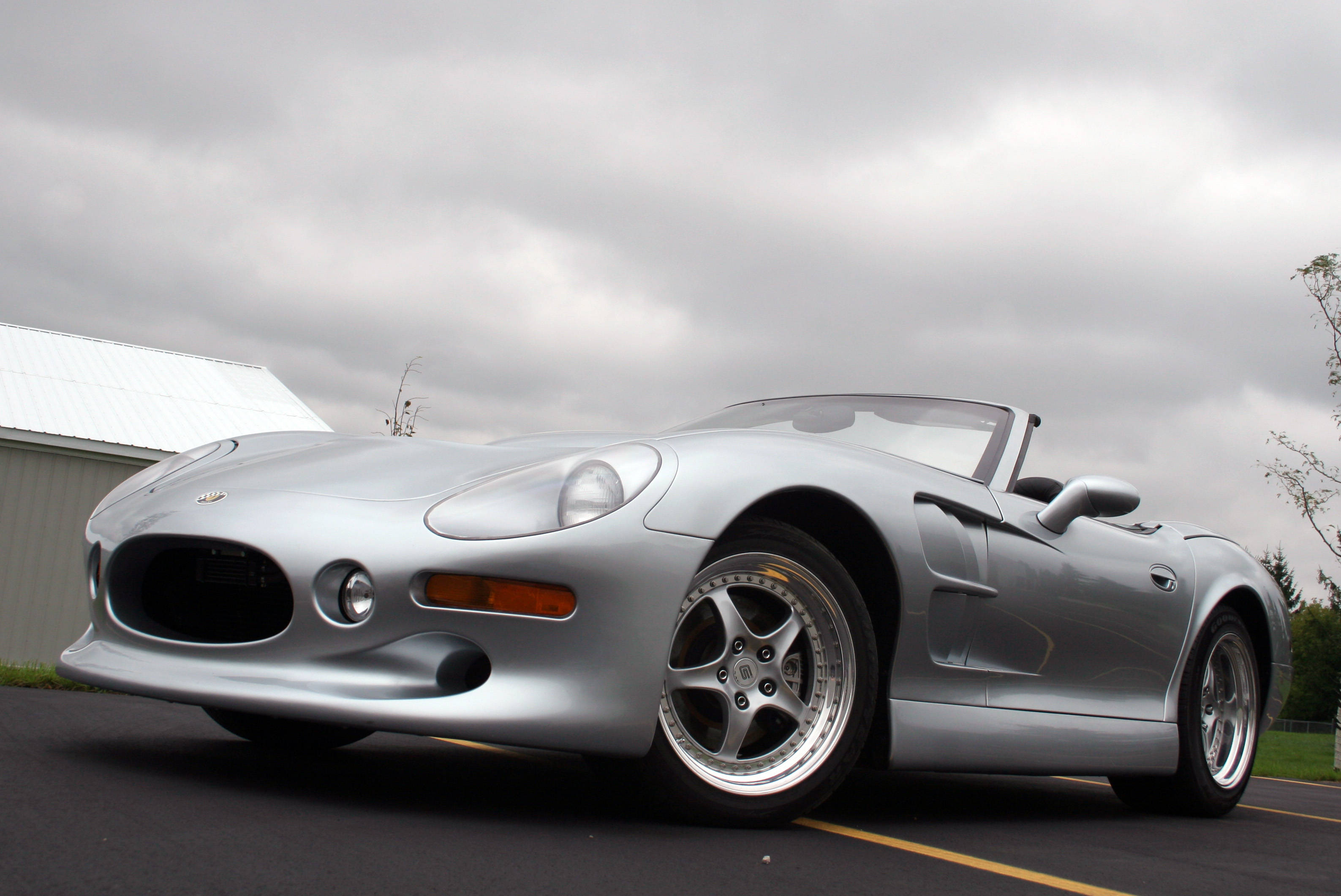 shelby iPhone 桌布 壁紙 — 一架時尚的銀色敞篷跑車停在瀝青路上，天空多雲，背景有穀倉。