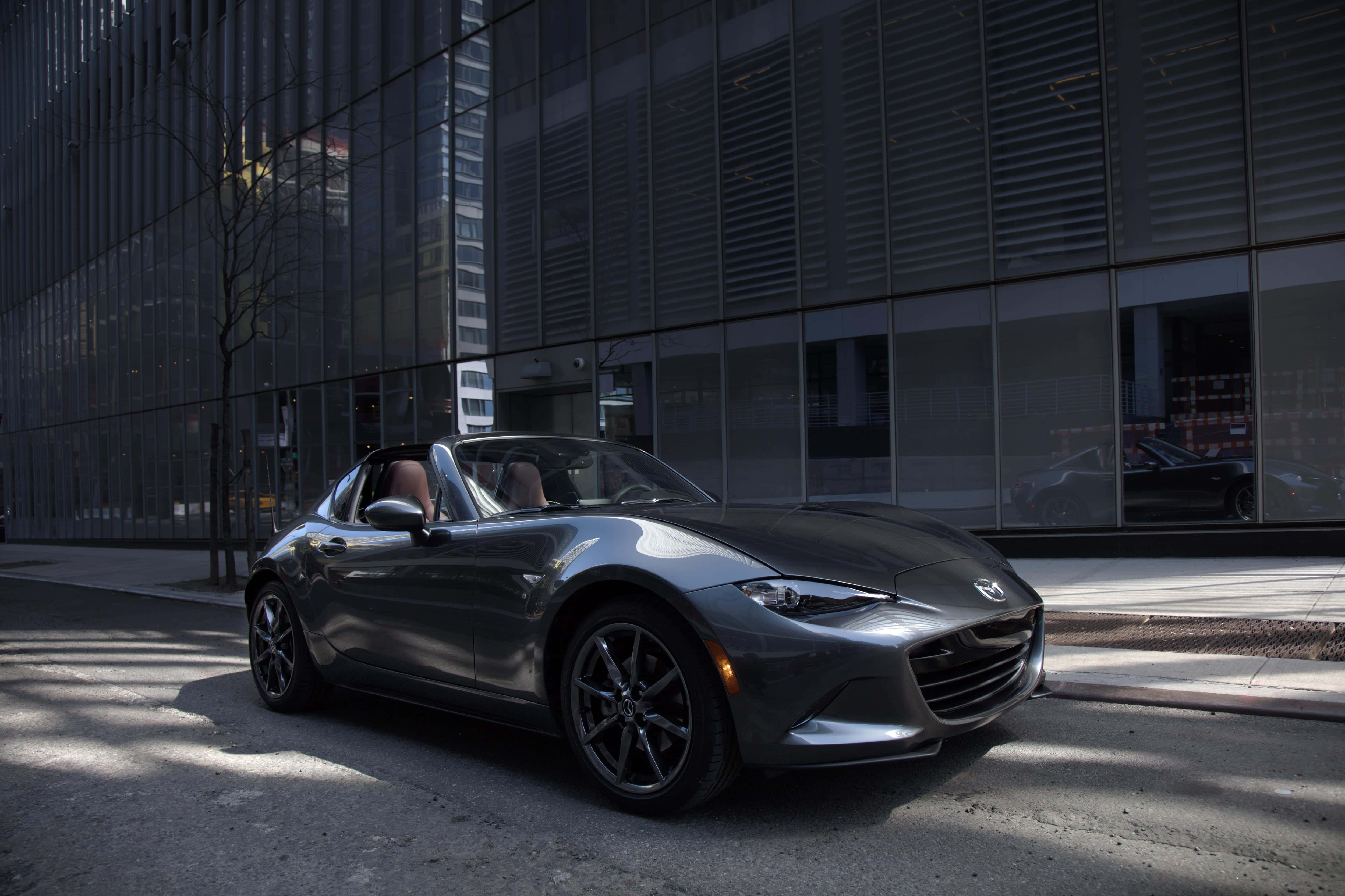 Miata 桌布 壁紙 — 一架時尚的銀色敞篷跑車停放在城市的街道上，旁邊是一座玻璃建築，光禿禿的樹木投下陰影。