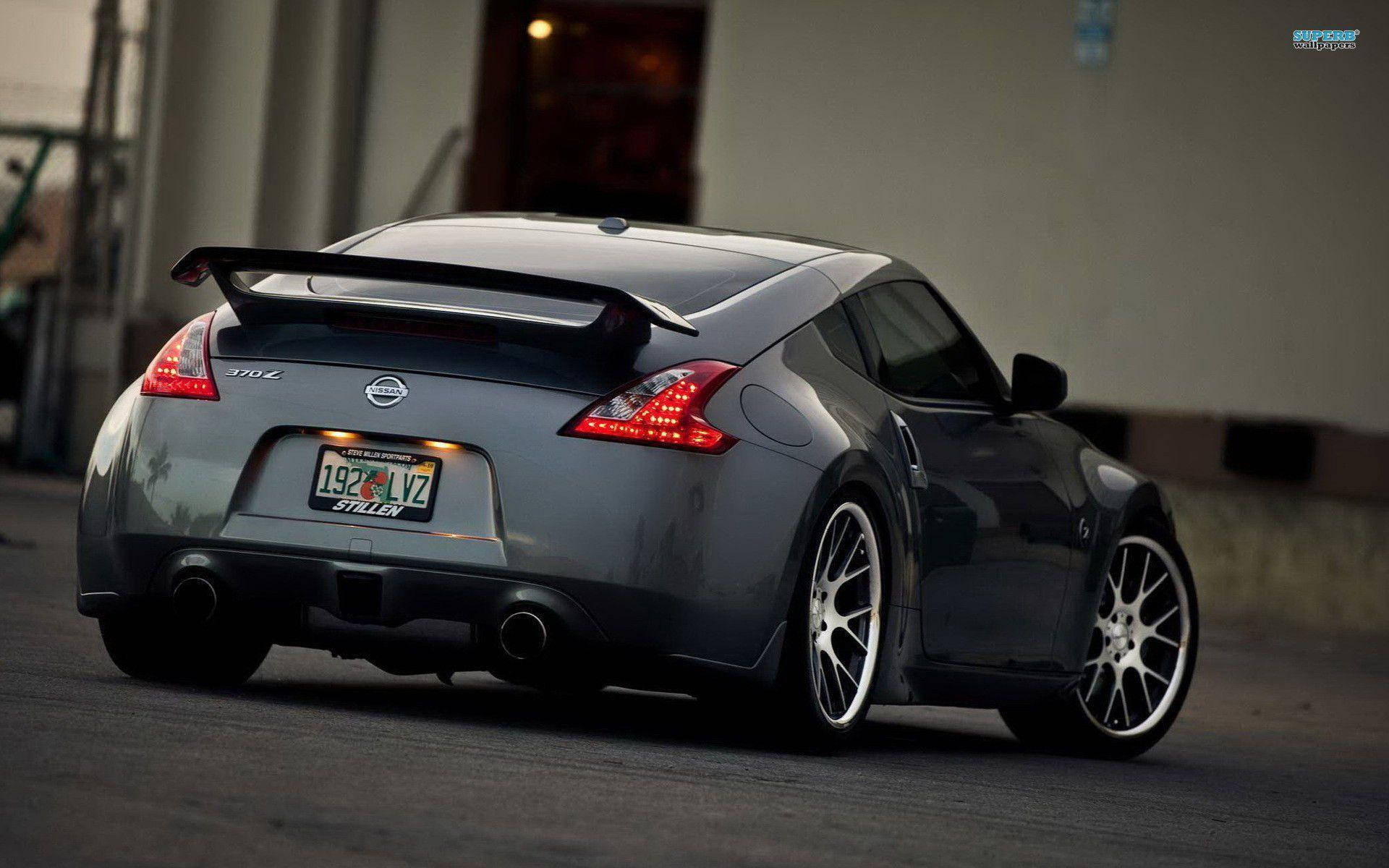 Nissan 370z 桌布 壁紙 — 一架時尚的銀色日產370Z跑車，配備黑色尾翼和訂製輪圈，停泊在燈光昏暗的城市街道上。