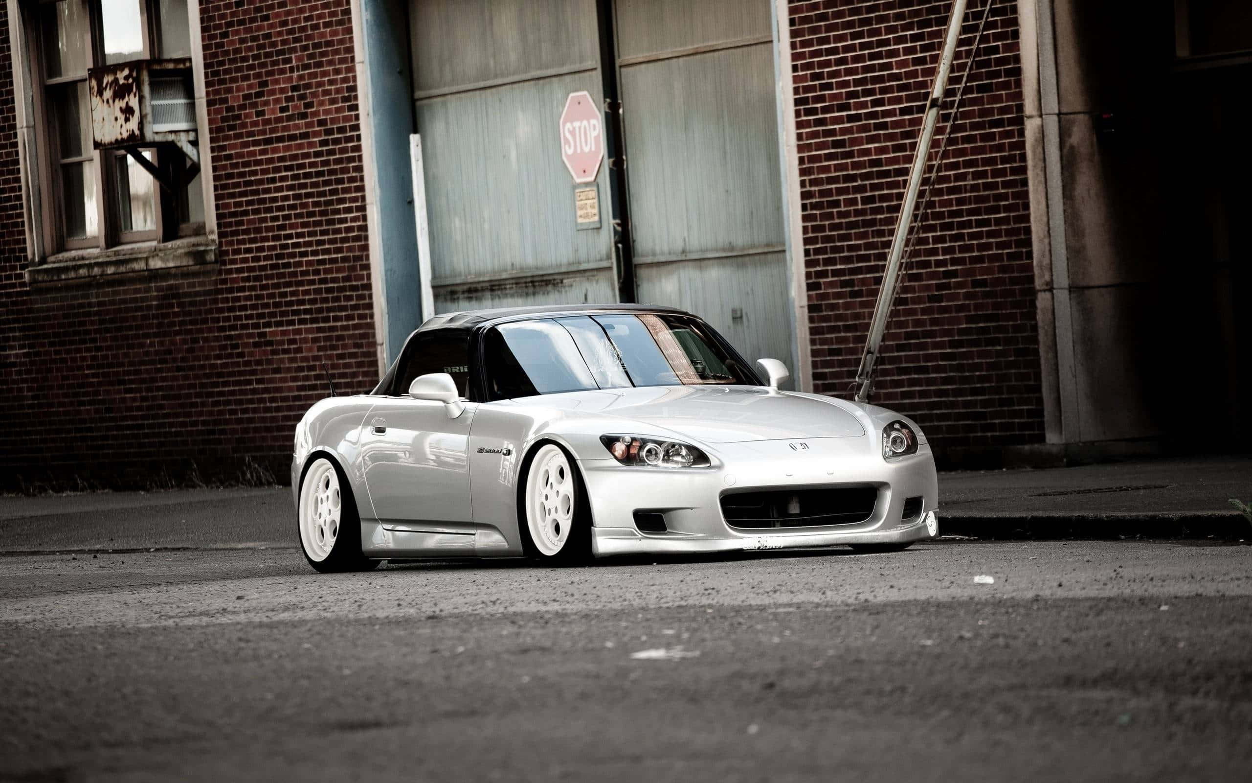 Honda S2000 桌布 壁紙 — 一架時尚的銀色本田S2000跑車停放在一個風化的磚砌倉庫前，旁邊有一個停車標誌。