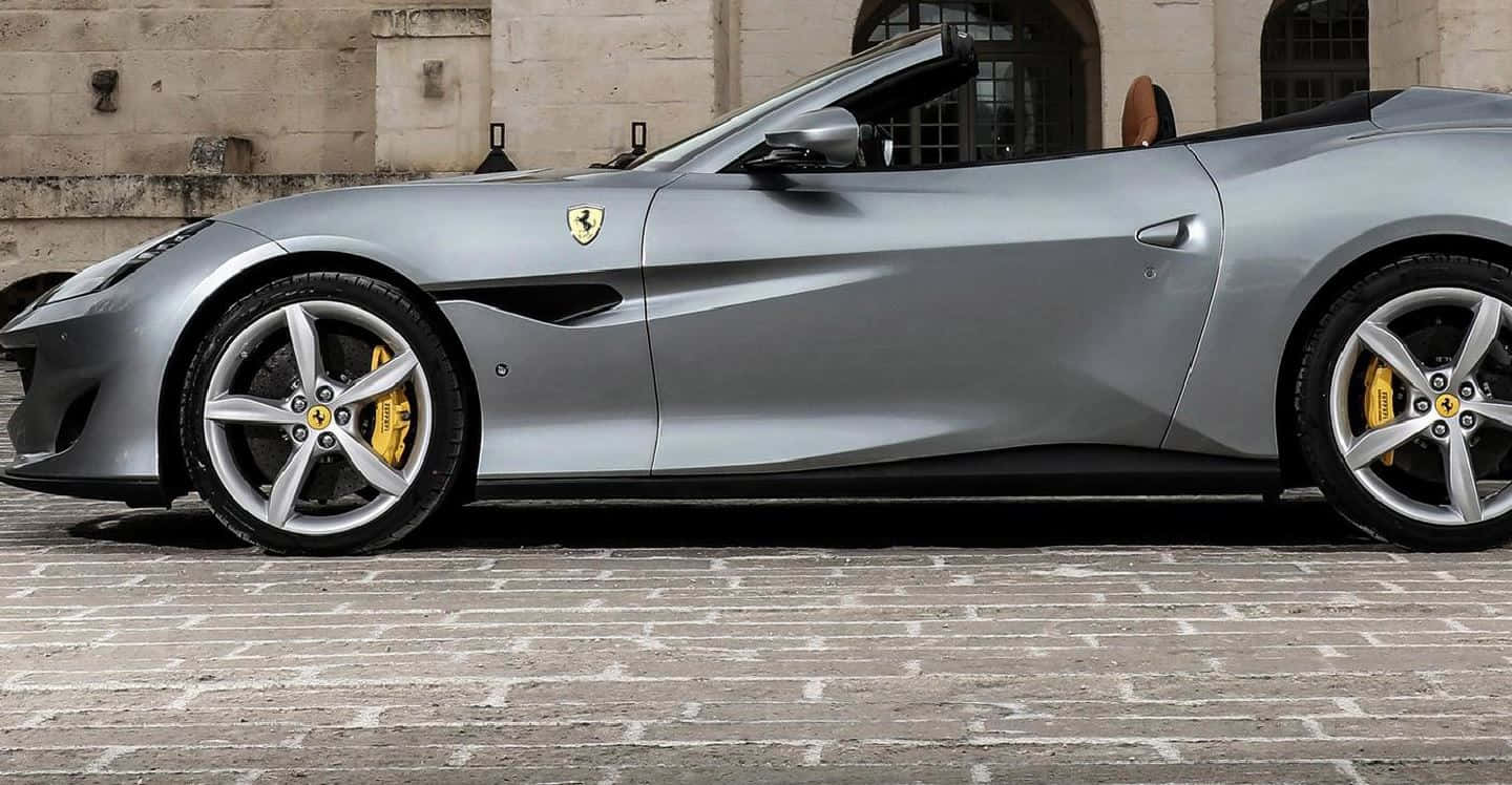 Ferrari Portofino 桌布 壁紙 — 一架時尚的銀色法拉利敞篷車，帶有黃色制動卡鉗，停泊在石砌建築旁的鵝卵石街道上。