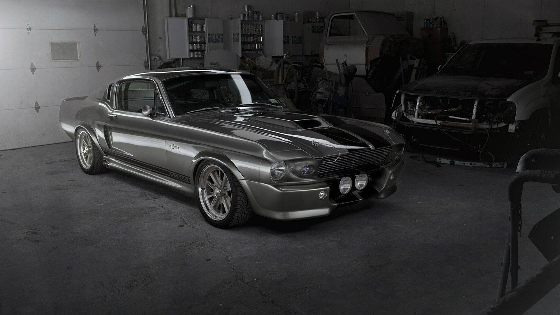 mustang HD 桌布 壁紙 — 一架時尚的銀色經典肌肉車帶有黑色賽車條紋，顯眼地停放在燈光昏暗的車庫中，周圍是汽車零件和另一輛正在維修的車輛。
