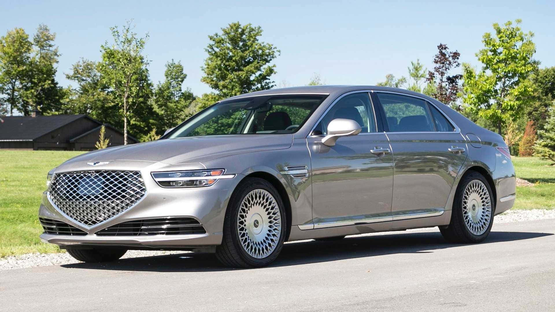 Genesis G90 桌布 壁紙 — 一架時尚的銀色豪華轎車停泊在鋪砌的道路上，背景有綠樹和房屋，天空清澈湛藍。
