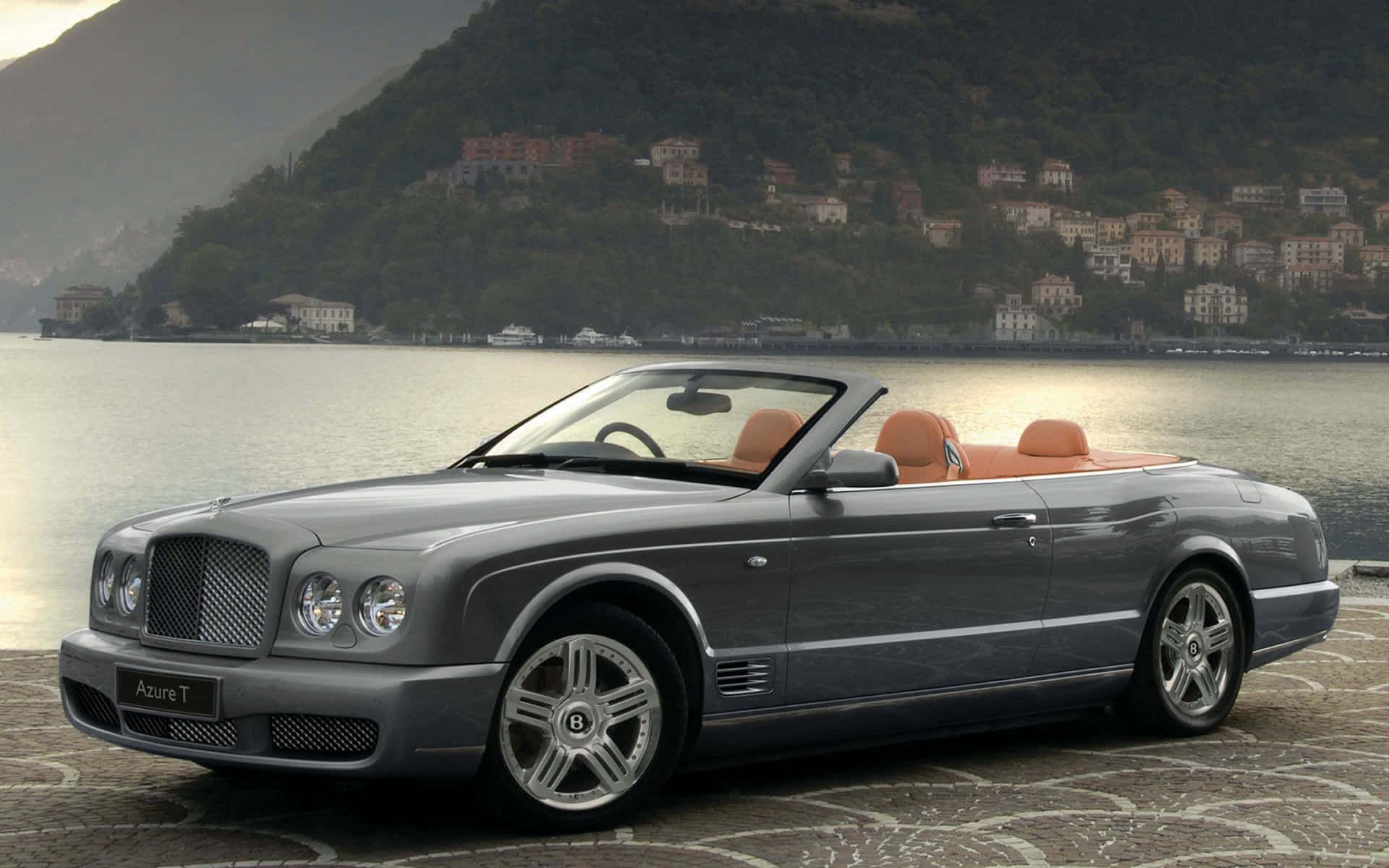 Bentley Azure 桌布 壁紙 — 一架時尚的銀色賓利 Azure T 敞篷車，配備棕色真皮座椅，停泊在平靜湖泊旁的石碼頭上，背景是山坡村莊。