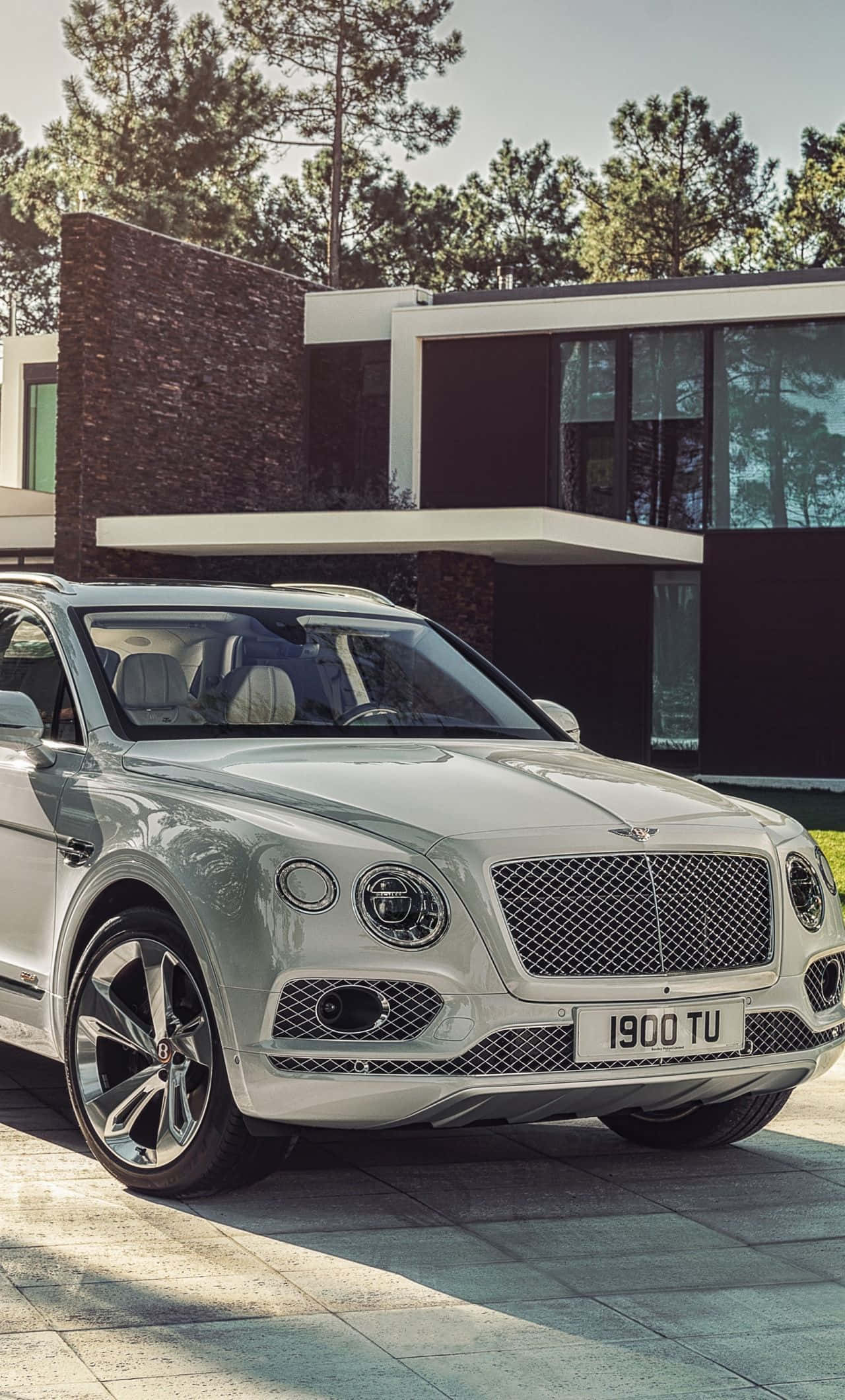 汽車 iPhone 壁紙 桌布 壁紙 — 一架時尚的銀色賓利Bentayga SUV停放在松樹環繞的現代房屋前的石頭庭院上。