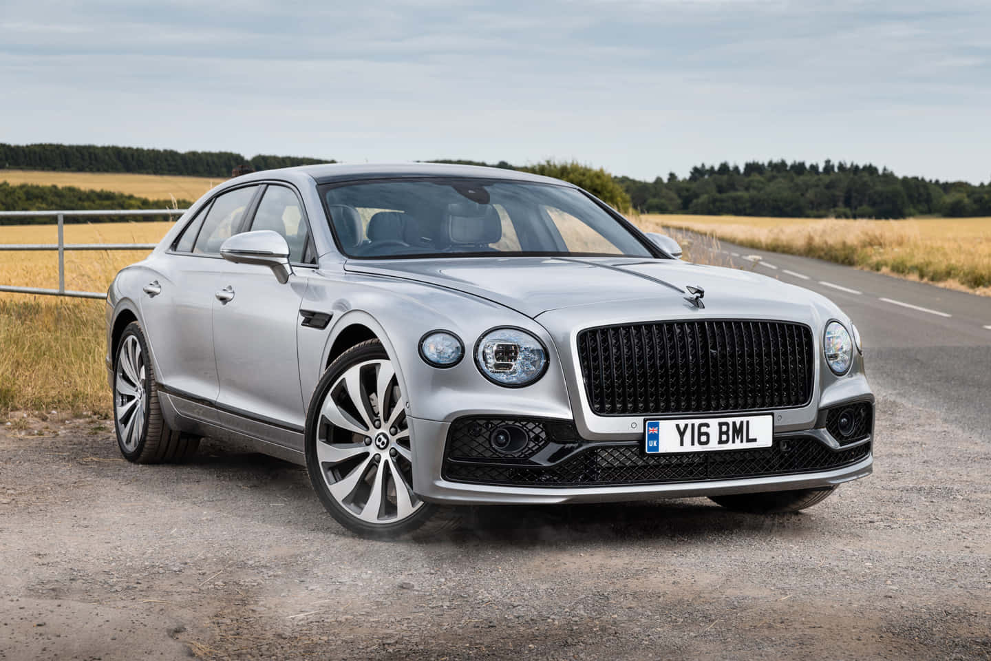 Bentley Flying Spur 桌布 壁紙 — 一架時尚的銀色賓利飛馳停泊在碎石路上，旁邊是金黃色的田野，天空多雲。