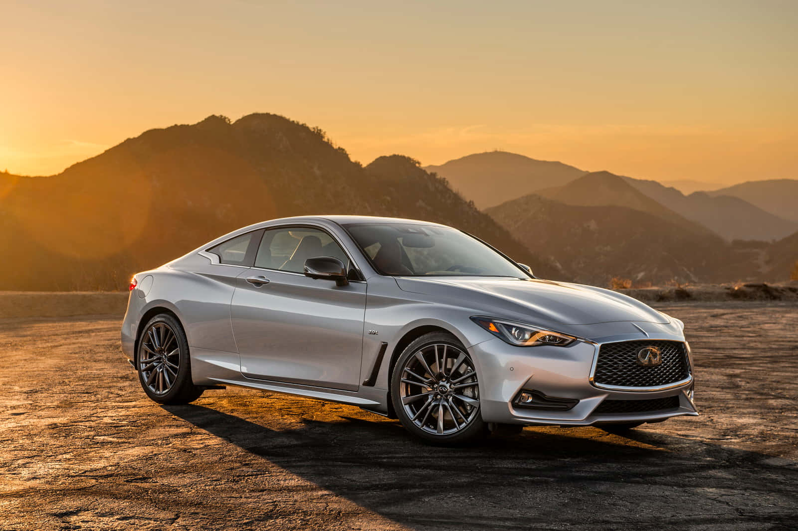 Infiniti Q60 桌布 壁紙 — 一架時尚的銀色跑車停放在裂開的路面上，背後是沐浴在金色日落光芒中的山脈。