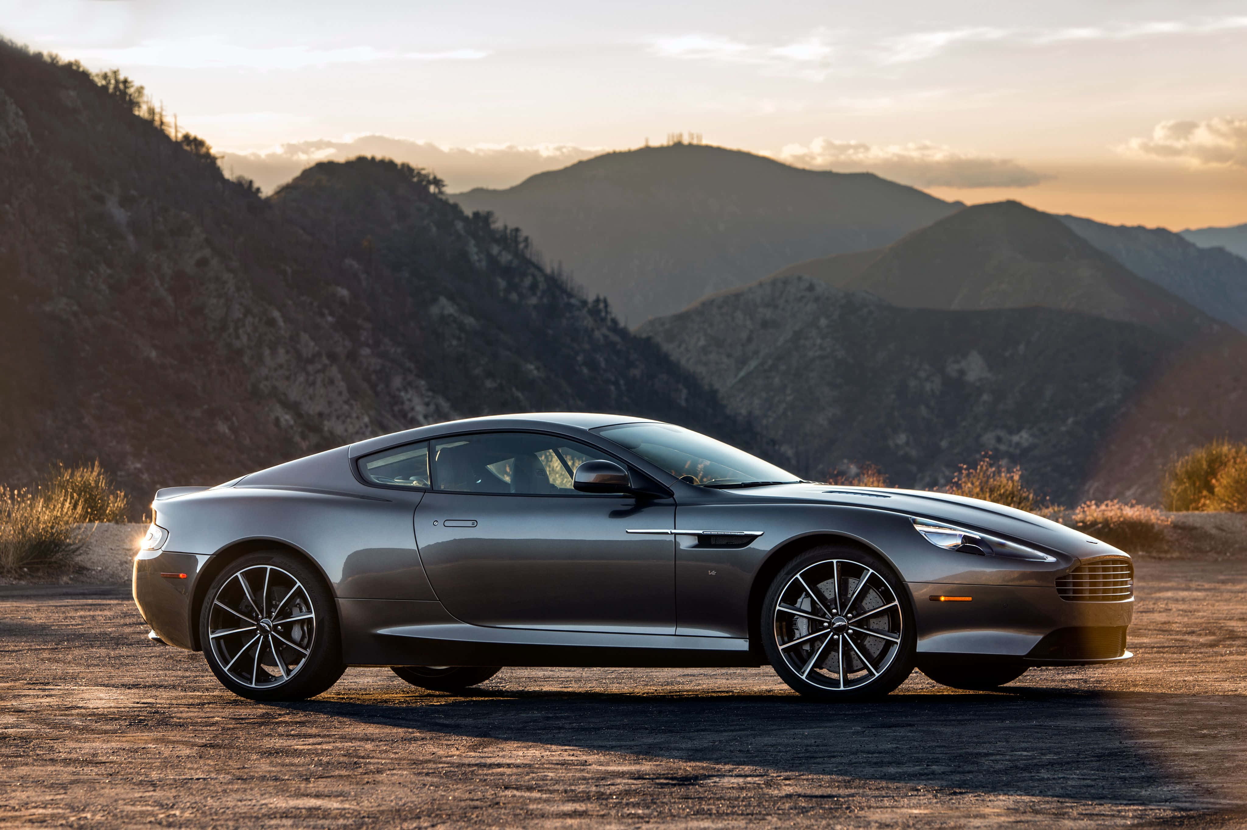 Aston Martin Db9 桌布 壁紙 — 一架時尚的銀色跑車停泊在塵土飛揚的道路上，山脈沐浴在金色的日落光芒中。