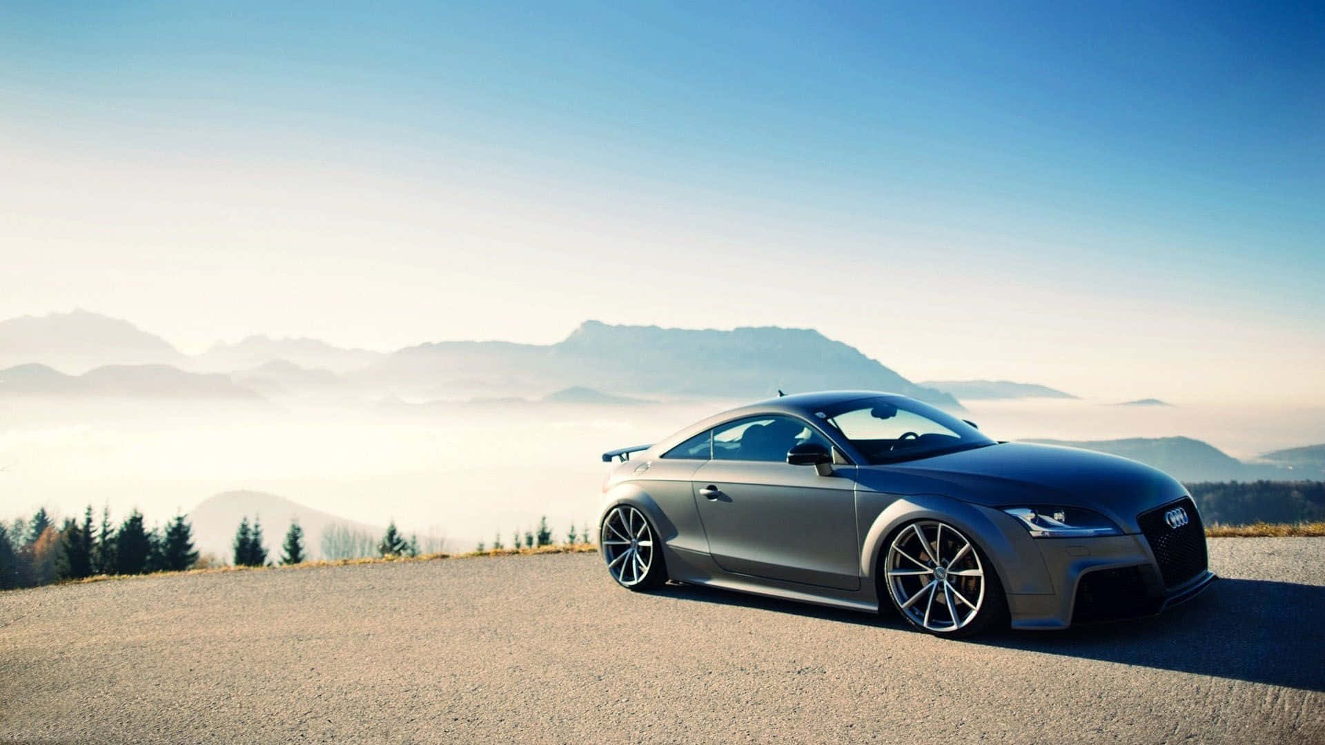 Audi Tt 桌布 壁紙 — 一架時尚的銀色跑車停泊在山路上，俯瞰著霧海和遠處的山峰，天空清澈湛藍。