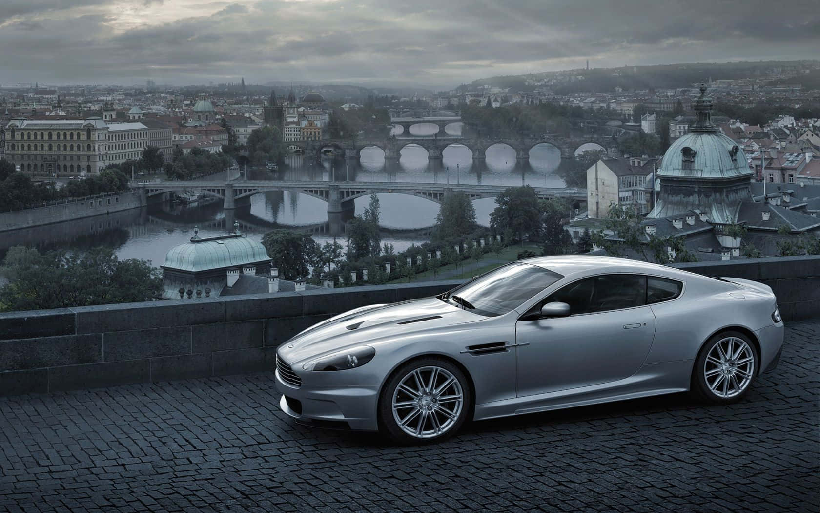 Aston Martin Vanquish 桌布 壁紙 — 一架時尚的銀色跑車停泊在鵝卵石觀景點上，霧氣瀰漫的河橋和歷史悠久的城市景觀在陰天之下。