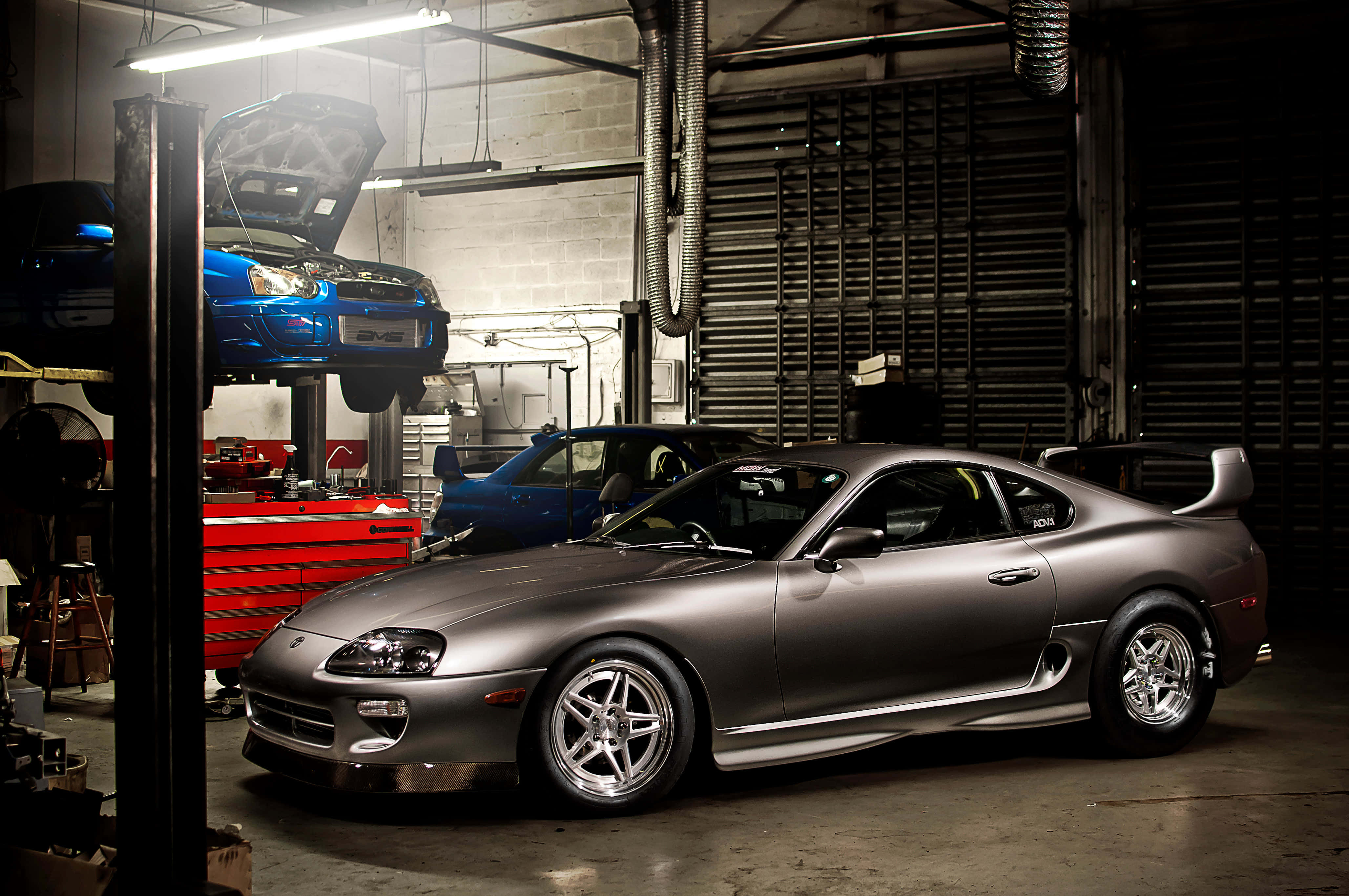 Supra 桌布 壁紙 — 一架時尚的銀色跑車在車庫燈光下閃閃發光，旁邊有一輛藍色汽車在升降機上，周圍是工具和設備。