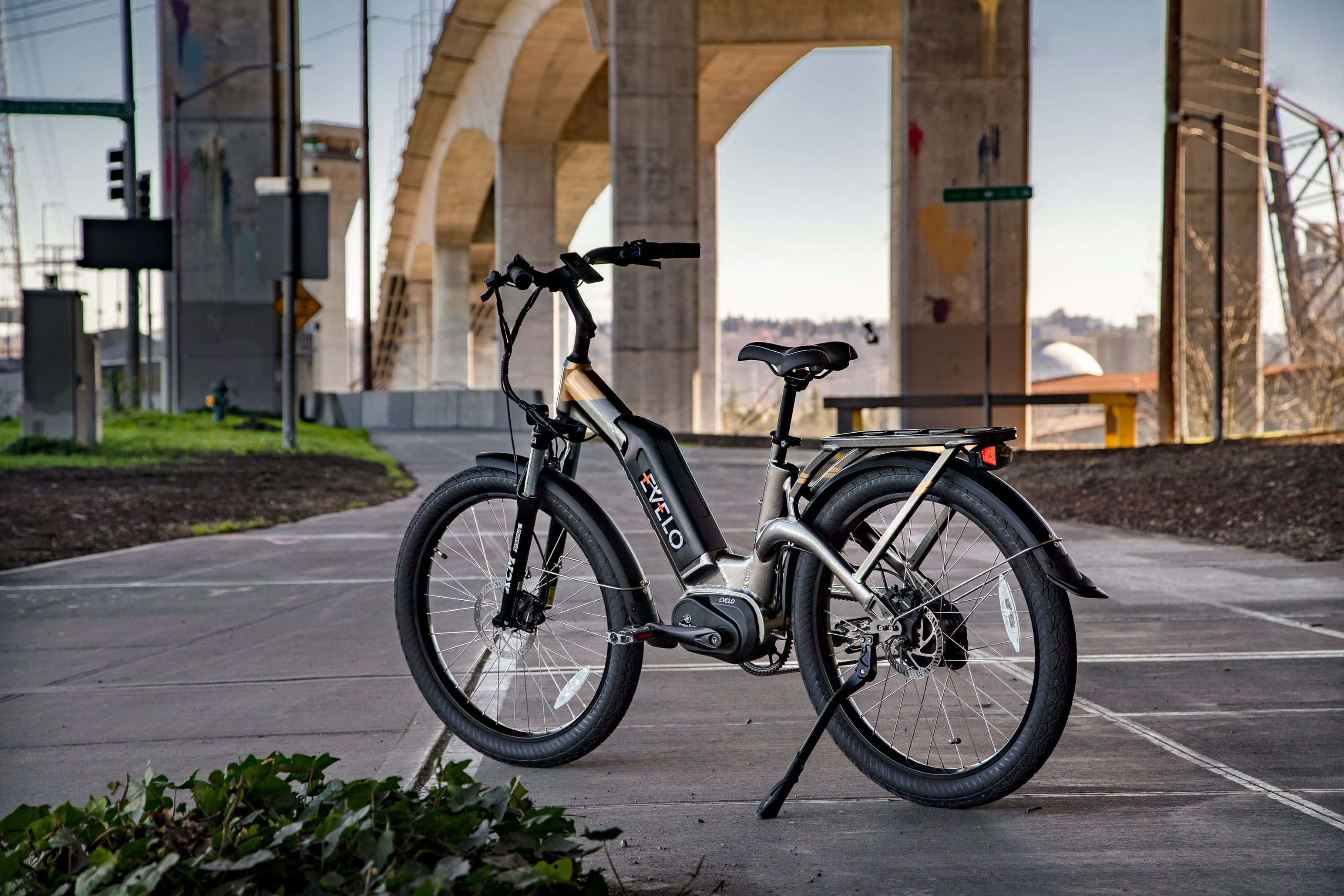Electric Bike 桌布 壁紙 — 一架時尚的銀色電動自行車停放在混凝土橋下，橋柱上有塗鴉，附近有綠化。