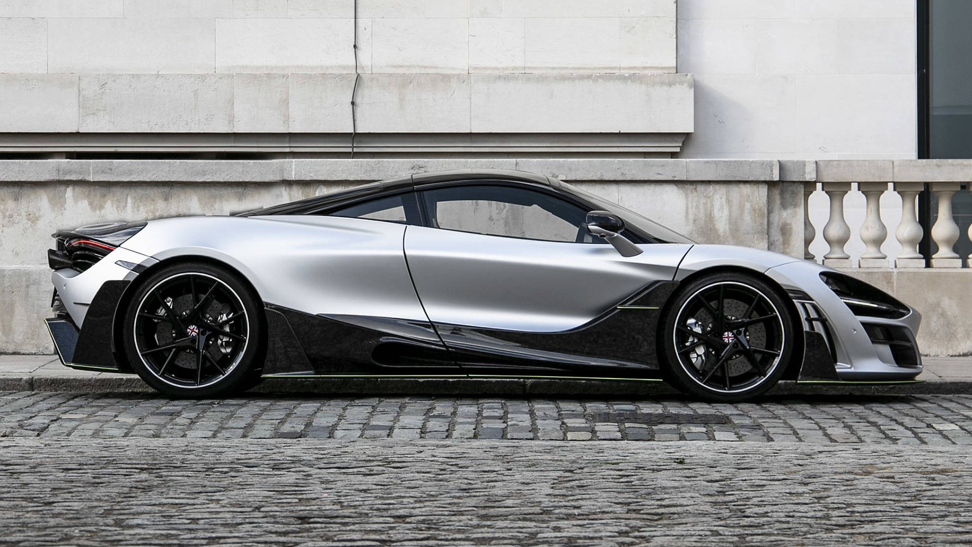 mclaren 720s 4K 桌布 壁紙 — 一架時尚的銀黑色McLaren 720S跑車停放在石砌建築旁的鵝卵石街道上。