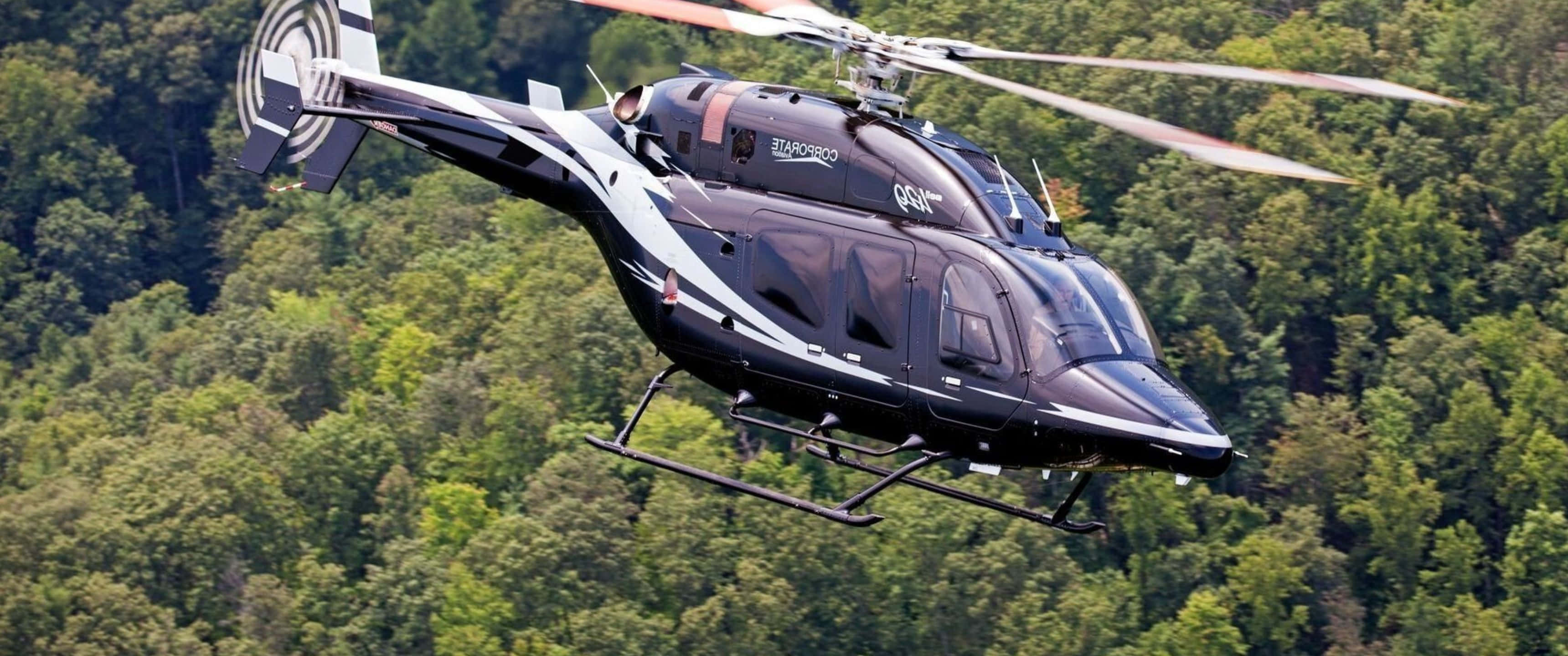 3440x1440p Helicopters 桌布 壁紙 — 一架時尚的黑白企業直升機在晴朗的日子低飛過茂密的綠色森林樹冠。