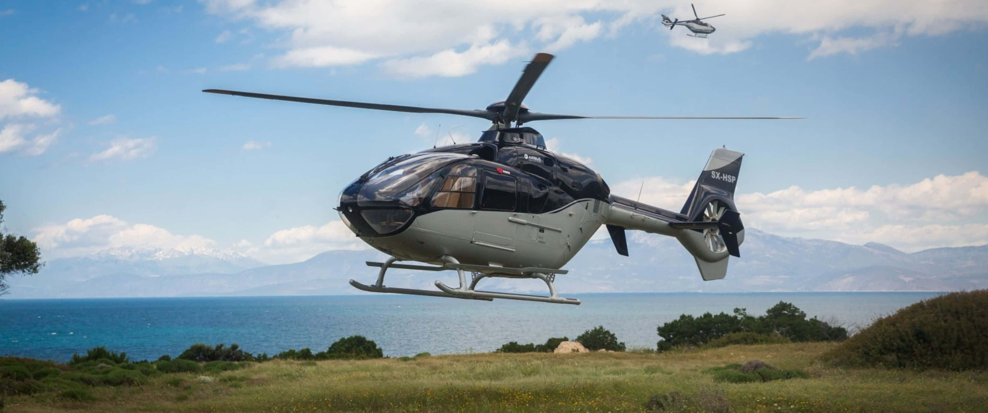 3440x1440p Helicopters 桌布 壁紙 — 一架時尚的黑白直升機盤旋在草叢海岸田野上空，另一架直升機在遠處明亮的藍天下飛行。