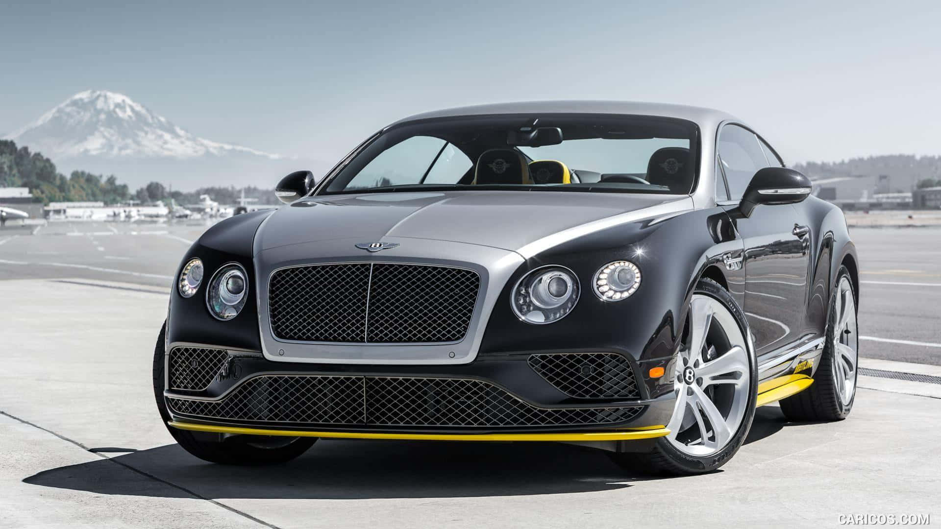 豪華車 桌布 壁紙 — 一架時尚的黑色 Bentley Continental GT 帶有黃色點綴，停在機場停機坪上，背景是白雪覆蓋的山峰。