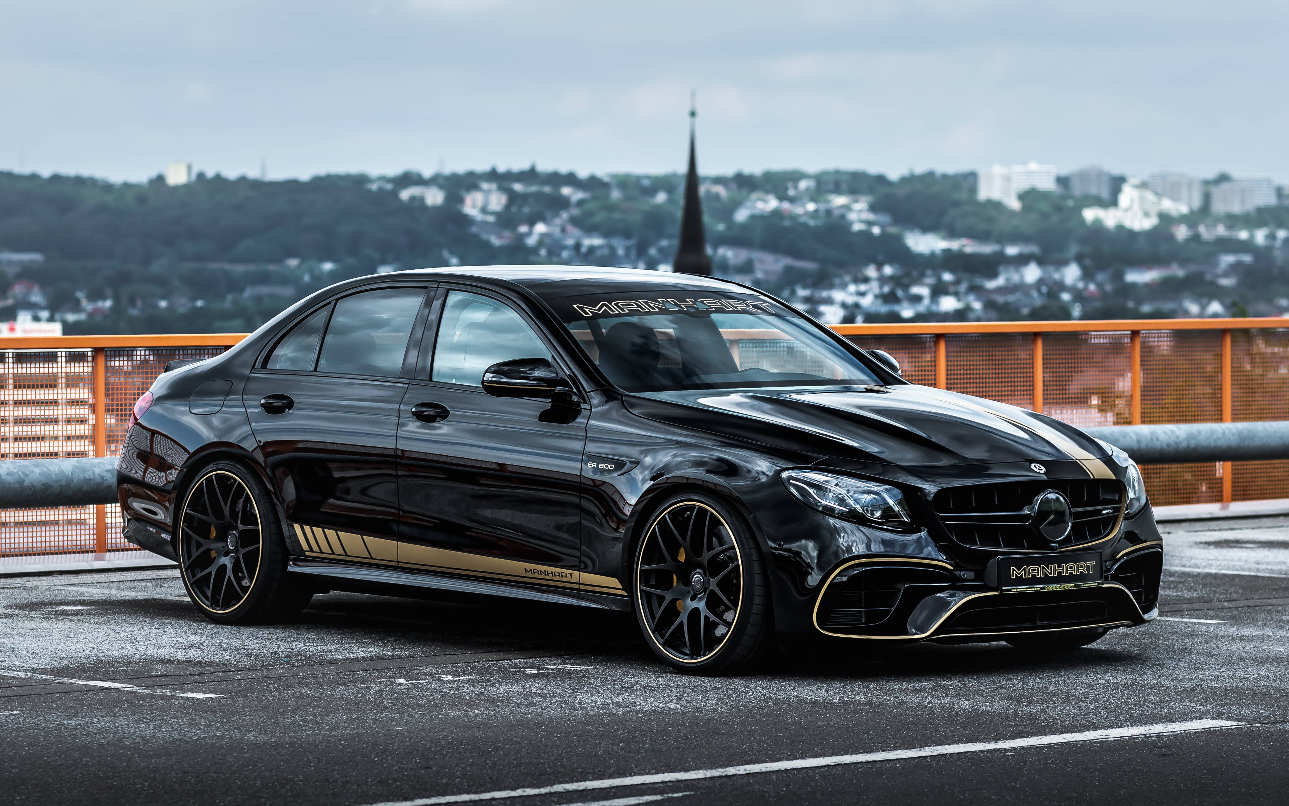 Amg 桌布 壁紙 — 一架時尚的黑色 Mercedes-AMG E63 S 帶有金色點綴，停泊在橋上，俯瞰陰天下的城市景觀。