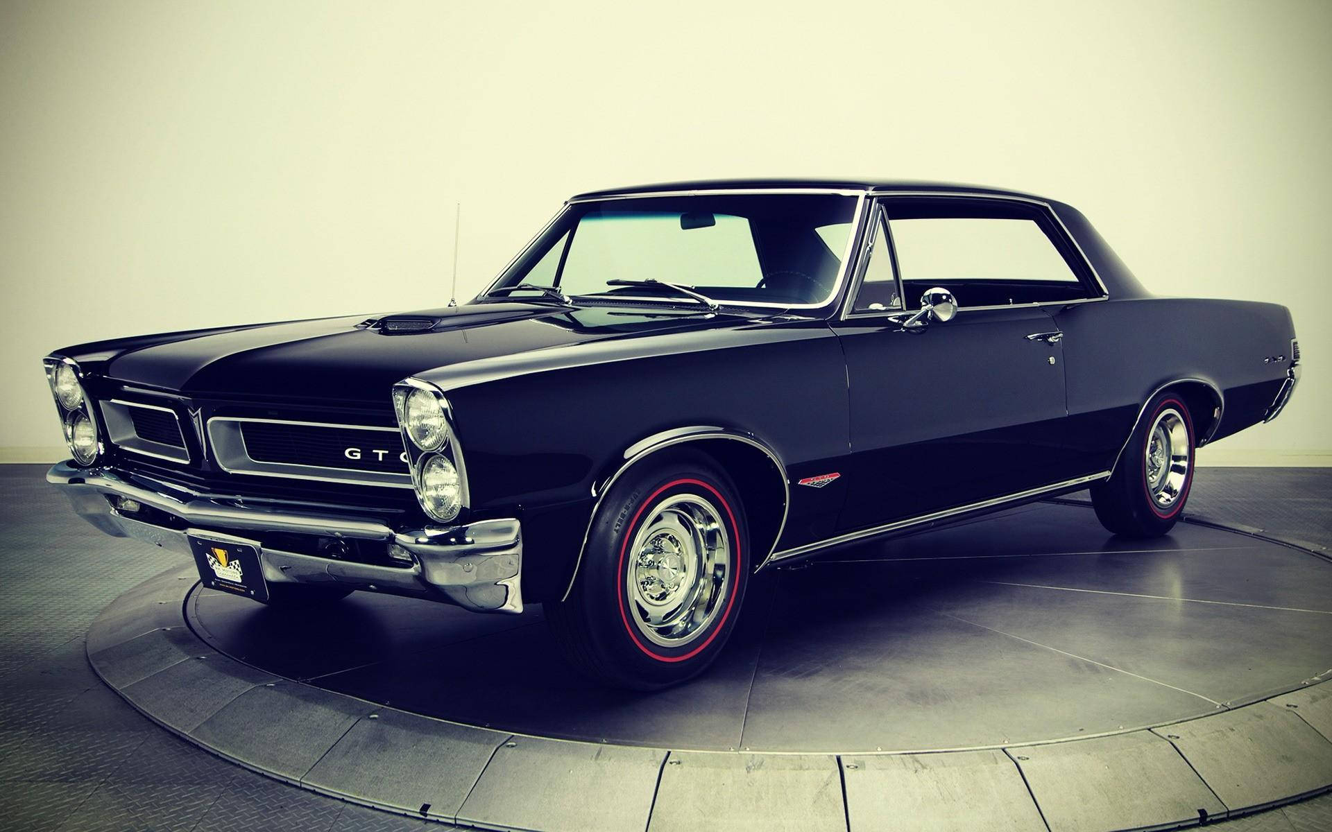 Pontiac 桌布 壁紙 — 一架時尚的黑色1960年代龐蒂亞克GTO肌肉車，配備紅線輪胎，在柔和的工作室燈光下，停放在圓形展廳旋轉台上。