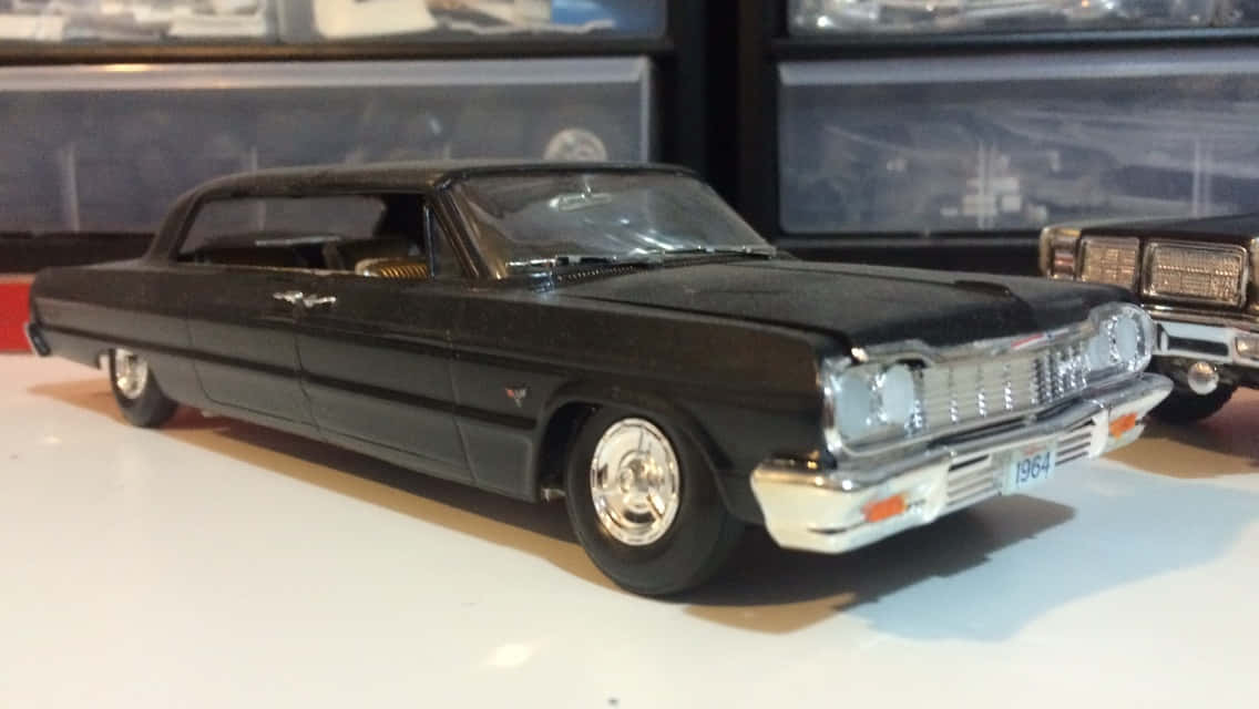 Car Model 桌布 壁紙 — 一架時尚的黑色1964年Cadillac模型車，帶有鍍鉻細節，放置在白色表面上，背景有其他模型。
