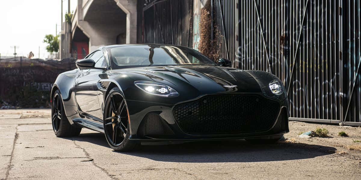 奧斯頓馬丁 桌布 壁紙 — 一架時尚的黑色Aston Martin跑車停泊在裂開的柏油路上，旁邊是佈滿塗鴉的金屬圍欄，在明亮的日光下。