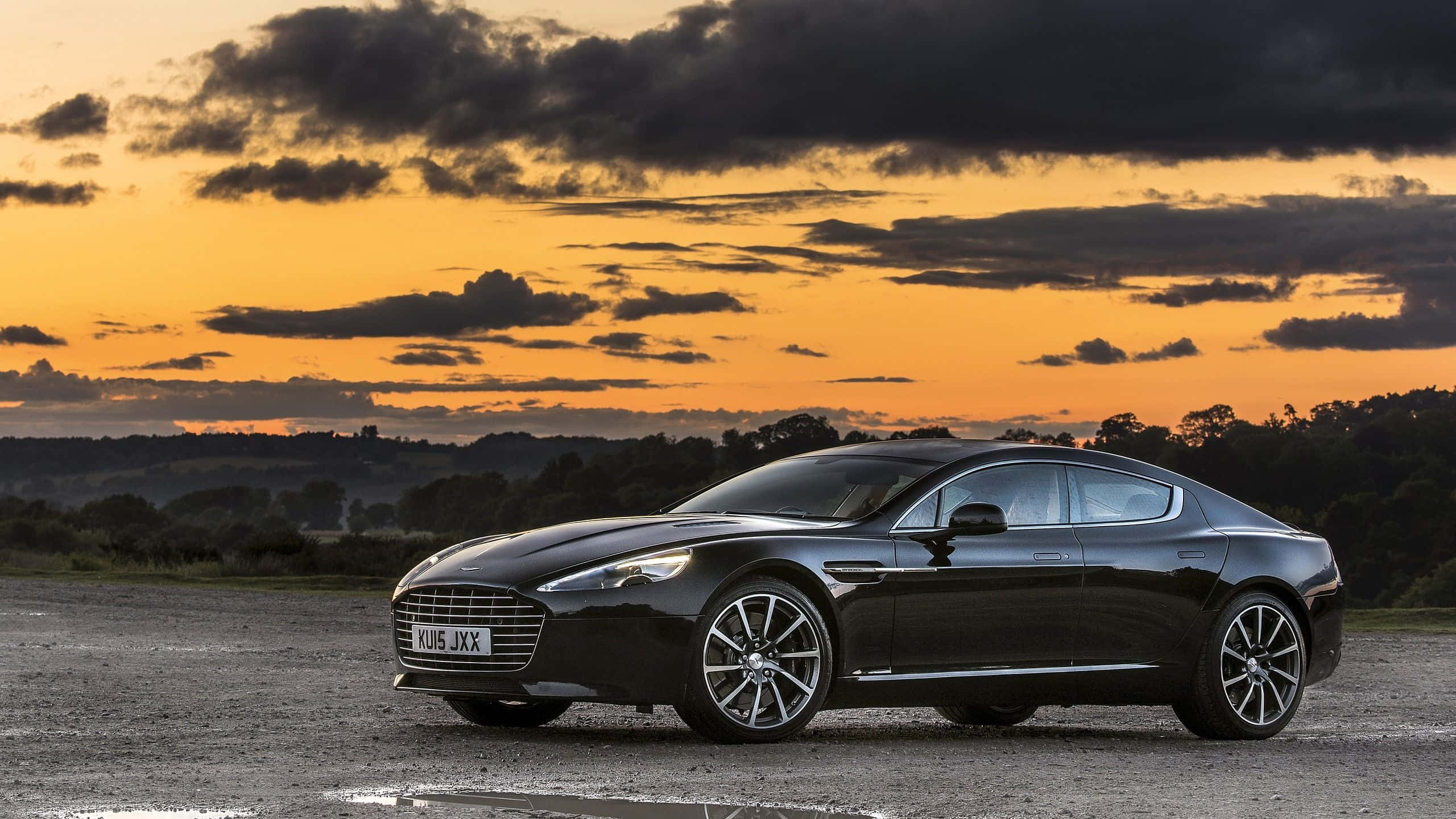 Aston Martin Rapide S 桌布 壁紙 — 一架時尚的黑色Aston Martin Rapide停放在碎石地上，背景是帶有橙色雲彩的戲劇性日落天空。