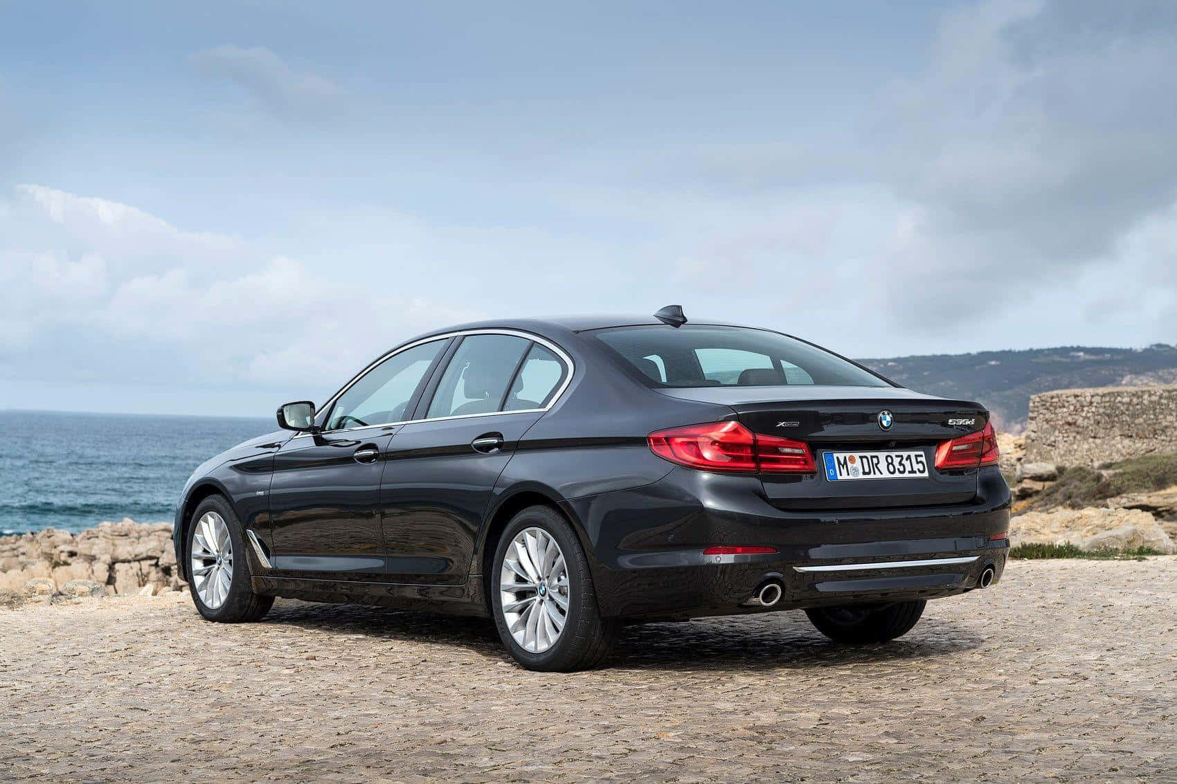 愛快羅密歐 桌布 壁紙 — 一架時尚的黑色BMW 5系列轎車停在鵝卵石懸崖上，俯瞰著多雲天空下的海洋。